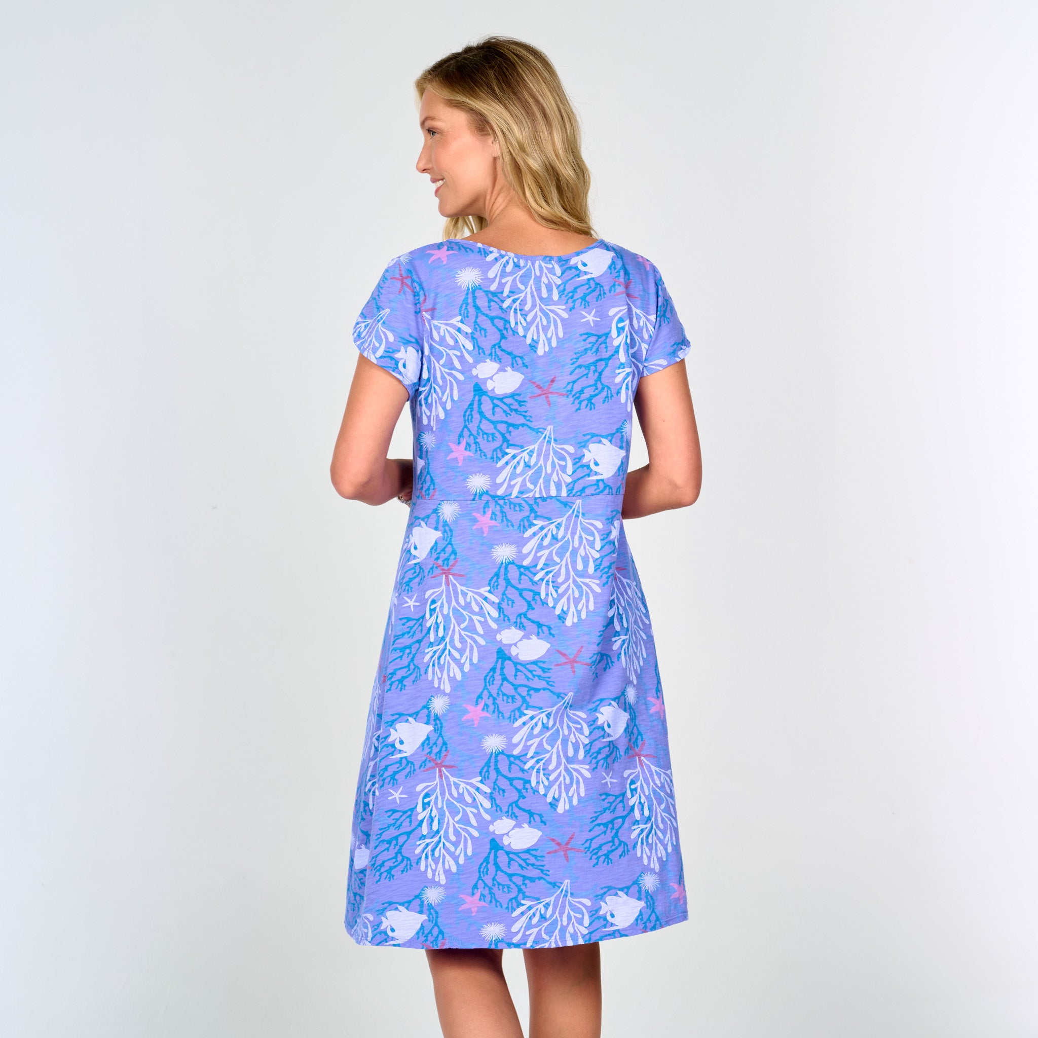 A woman stands with her back to the camera, wearing the Fresh Produce Clothes Coral Reef Amelia Dress—a knee-length, empire waist dress with short tulip sleeves, crafted from blue fabric decorated with a white coral and starfish pattern—set against a plain light background.