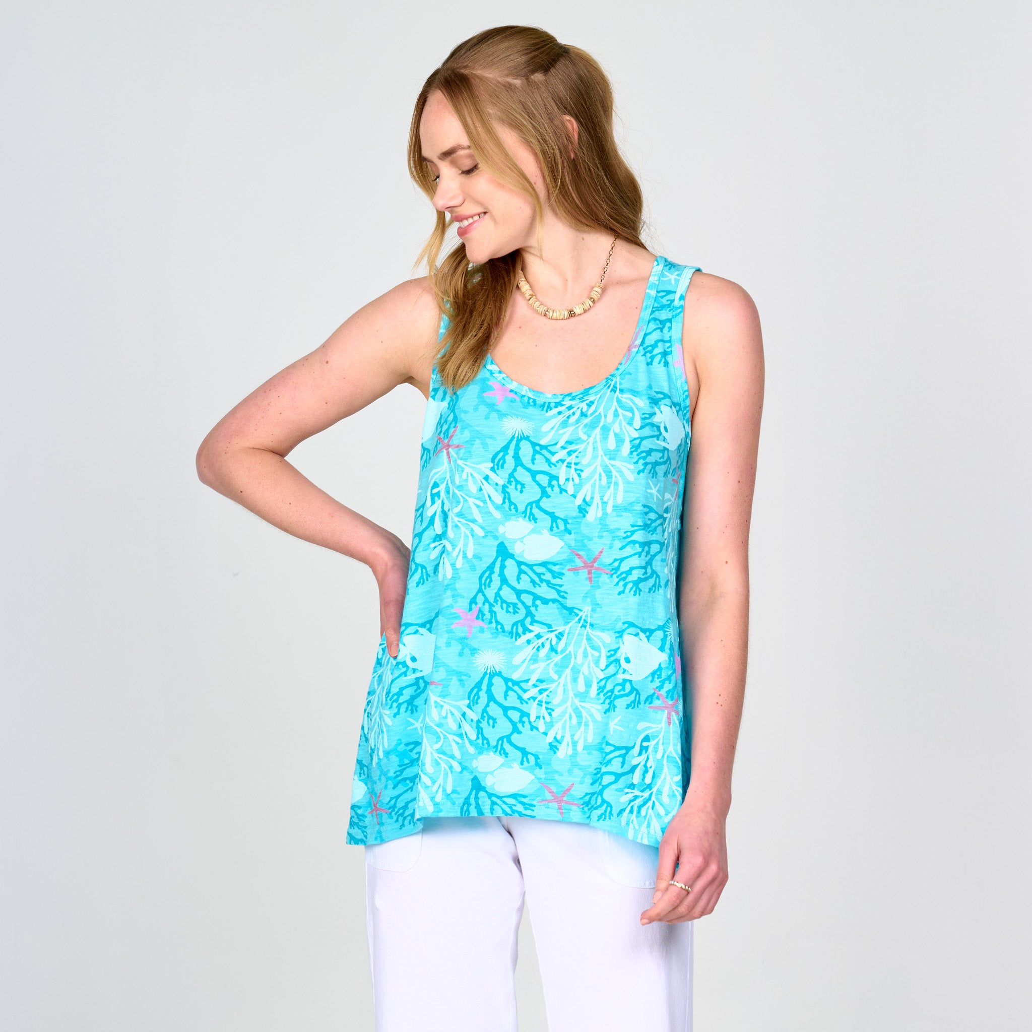 A woman smiles and gazes to her left against a plain light background, wearing the Fresh Produce Clothes Coral Reef Crossback Tank in a sleeveless turquoise, sea-inspired pattern paired with white pants.