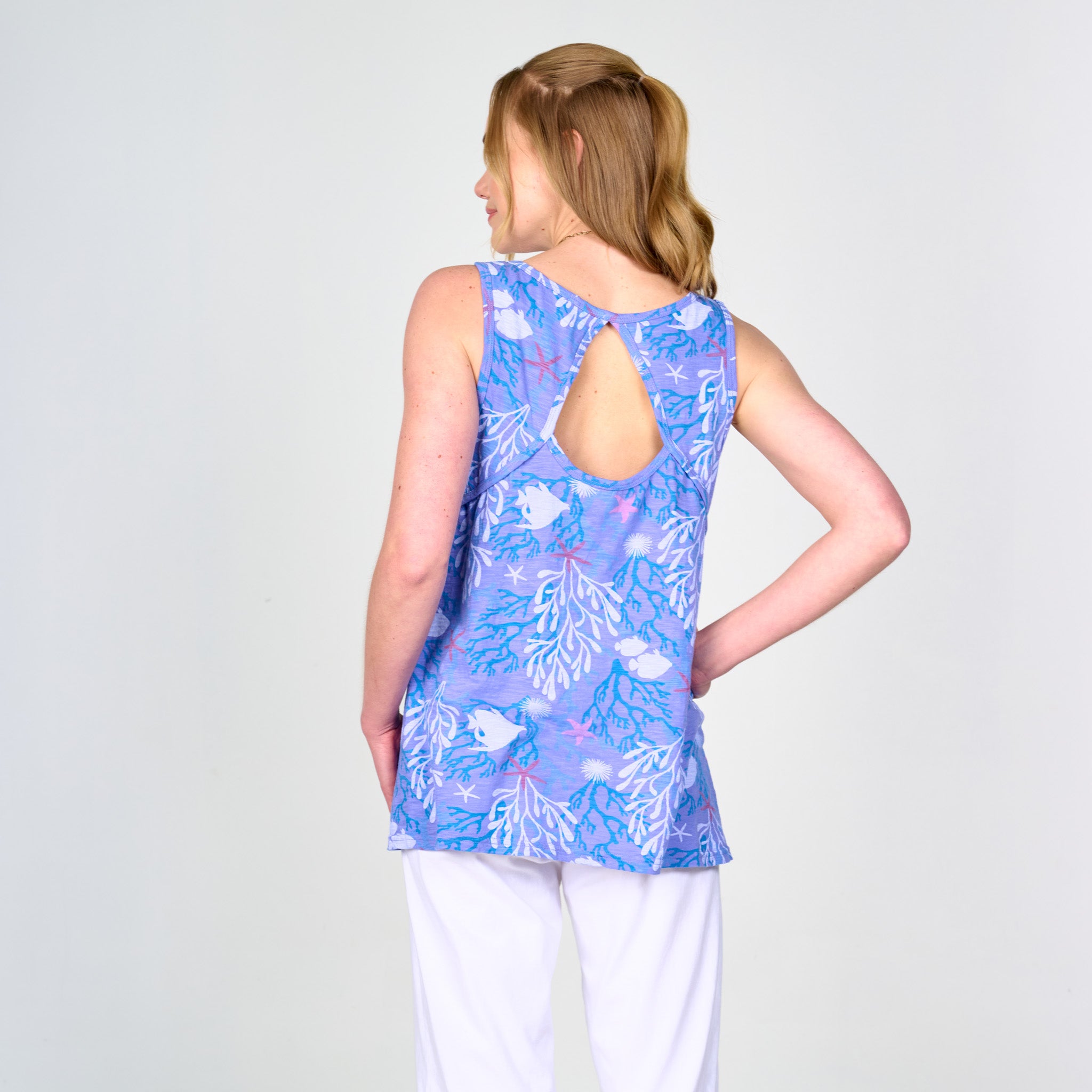 A woman stands facing away, wearing the Coral Reef Crossback Tank by Fresh Produce Clothes—a sporty top in a purple and blue pattern featuring a keyhole back detail—paired with white pants against a plain light background.