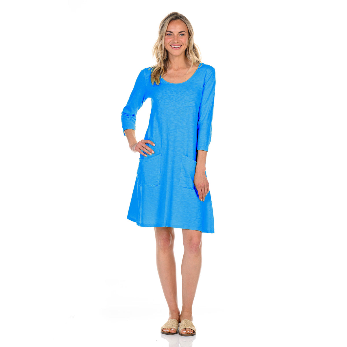 A woman stands smiling, wearing the bright blue Dalia Dress BFN SUF by Fresh Produce Clothes—a knee-length cotton slub dress featuring three-quarter sleeves and front pockets, proudly made in the USA. She has blonde hair and is wearing sandals. The background is plain white.