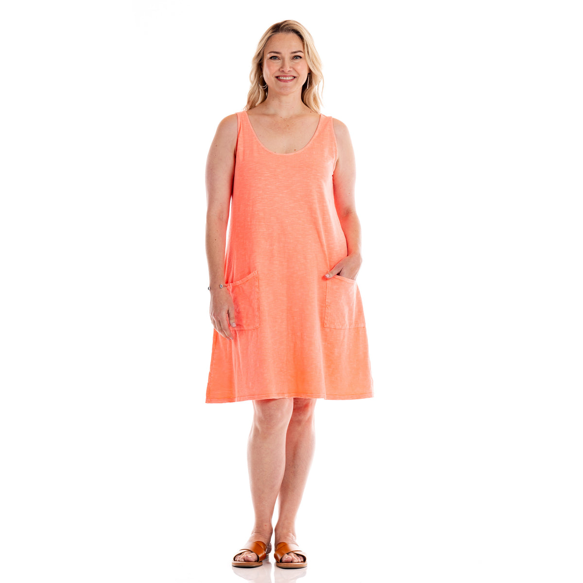 A smiling woman stands facing forward with one hand in the pocket of a sleeveless, coral-colored Drape Dress by Fresh Produce Clothes. This plus size cotton dress, featuring front pockets and brown sandals, is made in the USA and is available on clearance.