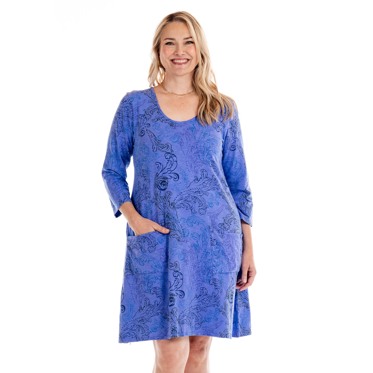 A woman smiles as she wears the Waverley Swirl Dalia Dress | Plus Size by Fresh Produce Clothes—a versatile, knee-length, long-sleeved blue dress featuring black paisley designs in the Waverley Swirl pattern and convenient front pockets—while standing against a white background.