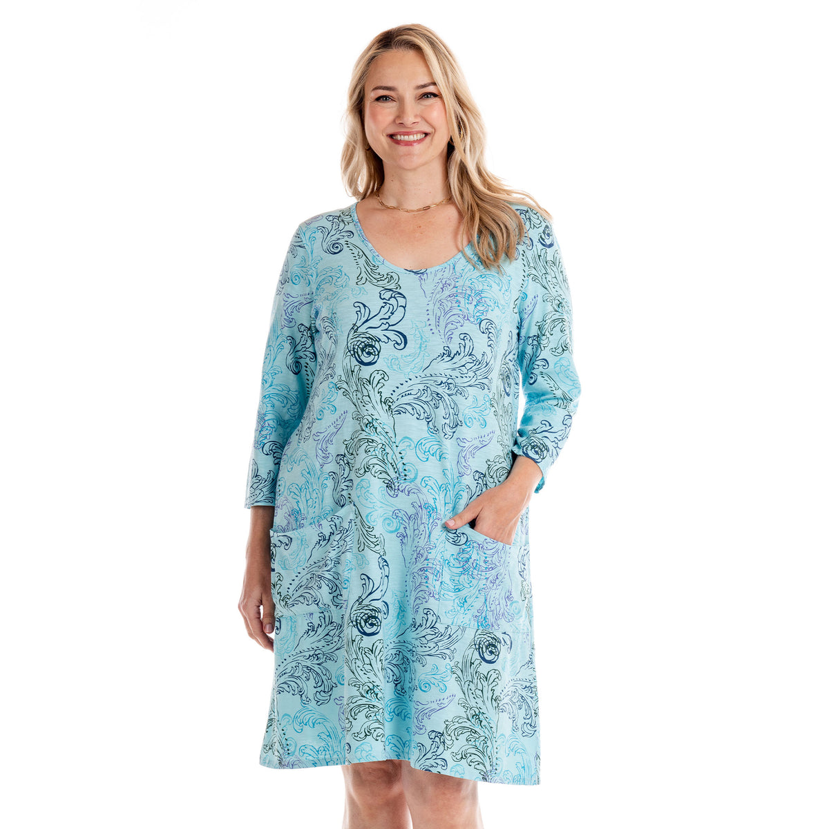 A woman smiles, wearing the Waverley Swirl Dalia Dress in light blue from Fresh Produce Clothes. This versatile, knee-length plus size cotton dress features long sleeves and a swirling paisley pattern, with one hand casually tucked in her pocket as she stands against a plain white background.