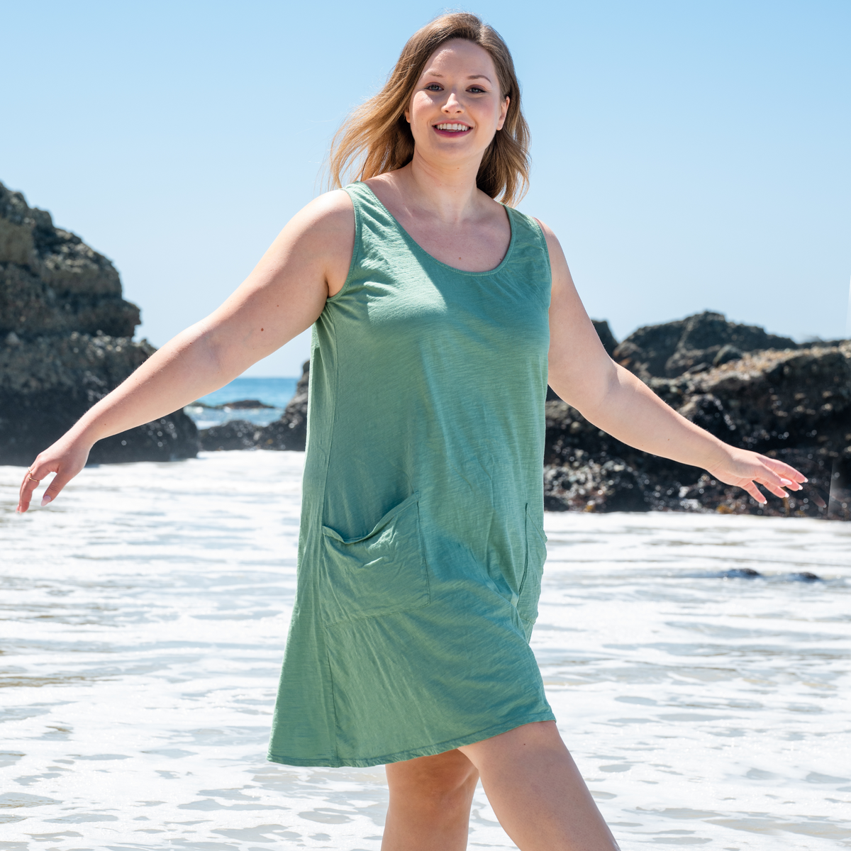 A woman wearing the Fresh Produce Clothes Drape Dress (Plus Size | Clearance), a sleeveless green cotton dress, strolls along the beach with a smile and arms outstretched. Rocky cliffs and ocean waves can be seen in the background beneath a clear blue sky.
