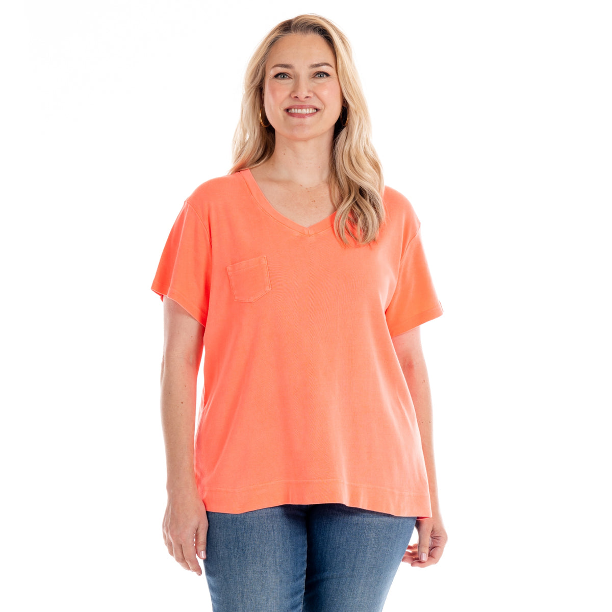 A woman with long blonde hair smiles while wearing the Fresh Produce Clothes Ribbed Logo Fem T in plus size Sunset Coral—a semi-fitted, ribbed cotton V-neck shirt with a chest pocket—paired with blue jeans and standing against a white background.