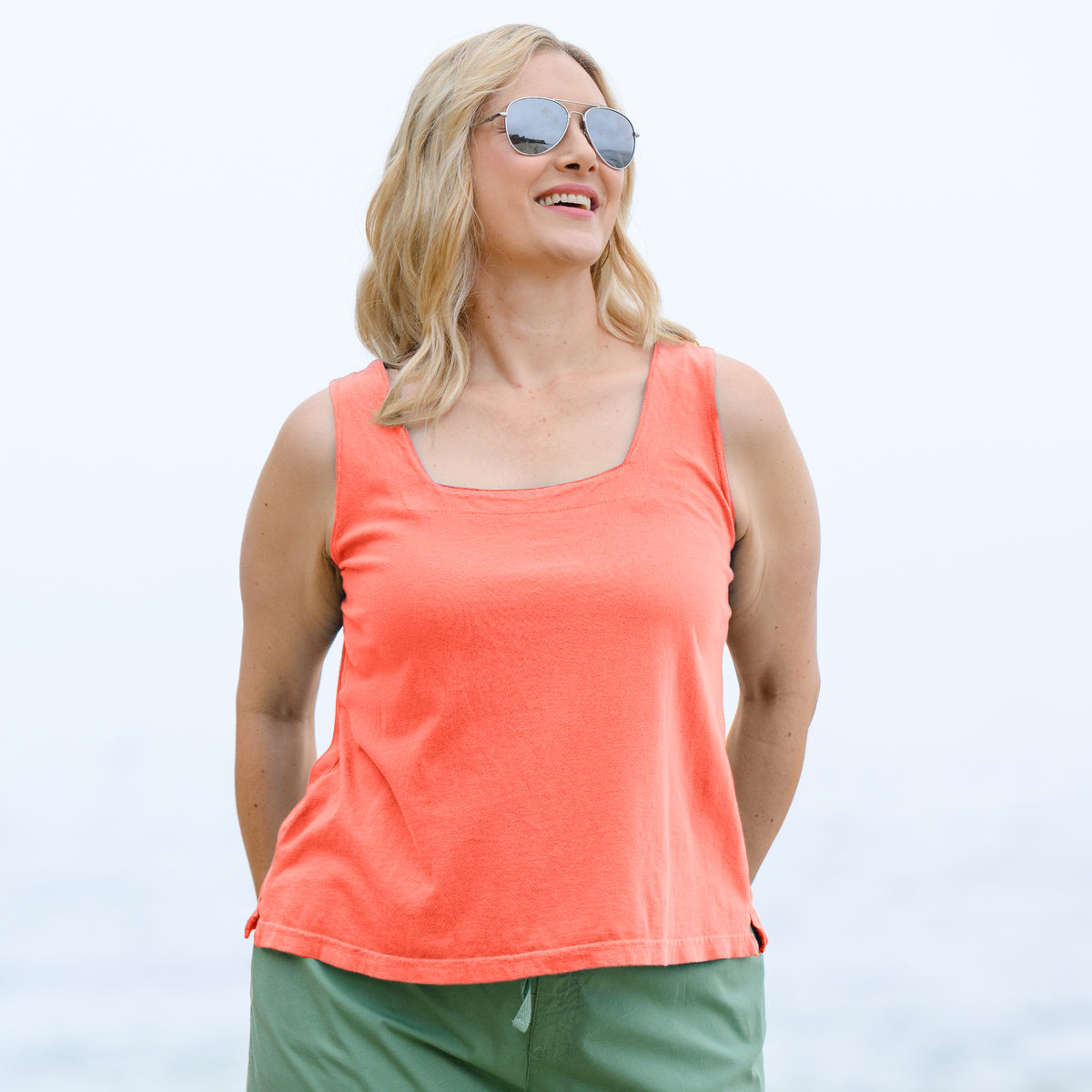 A woman with blonde hair in sunglasses and a Fresh Produce Clothes Boxy Tank | Plus Size Sunset Coral smiles, standing outdoors against a light, blurry background.
