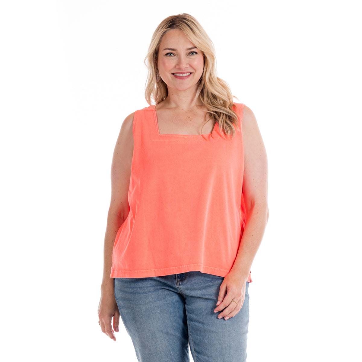 A smiling woman with long blonde hair wears the Fresh Produce Clothes Boxy Tank | Plus Size Sunset Coral, made from 100% cotton jersey, paired with blue jeans and standing against a white background.