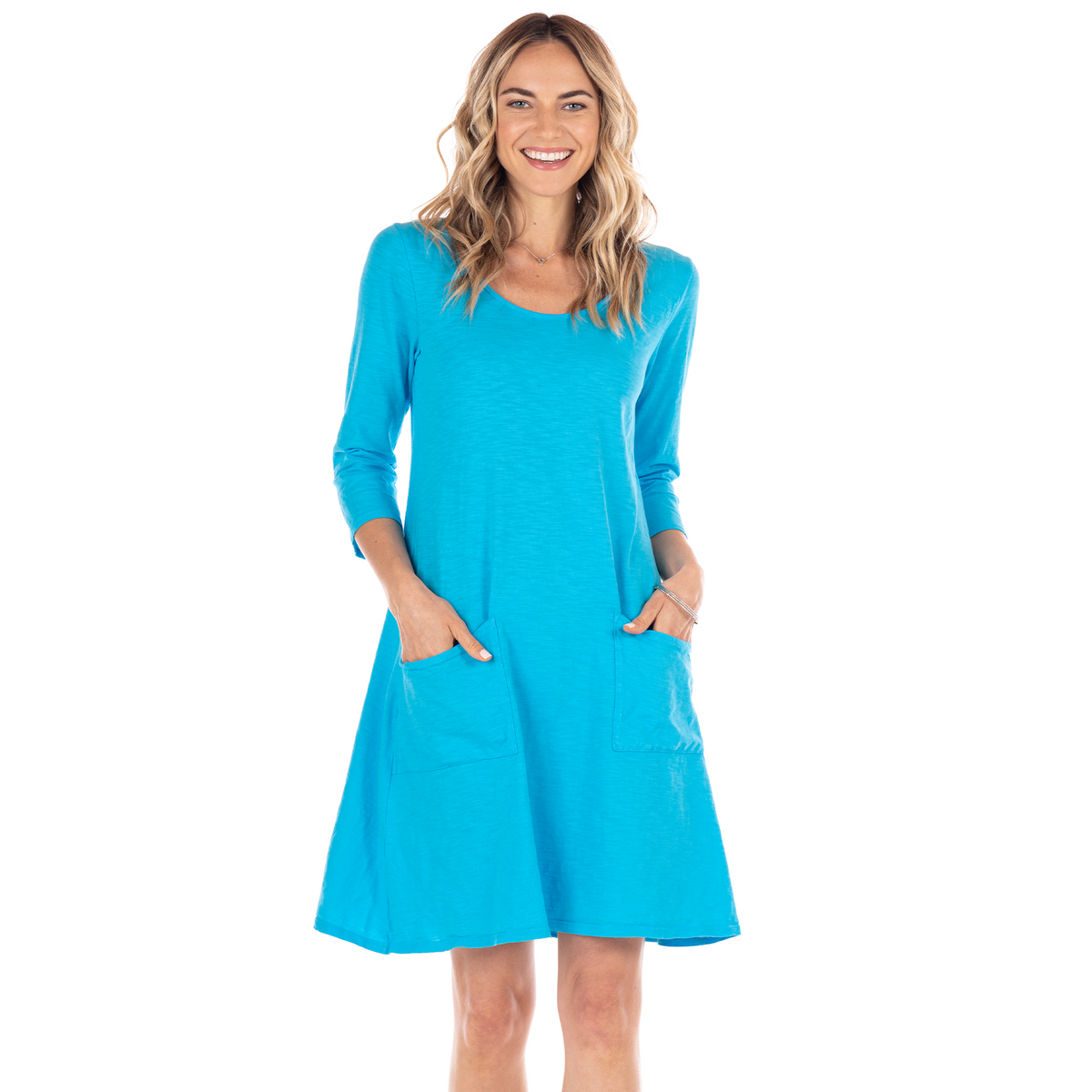 A woman smiles while wearing the bright blue, knee-length Dalia Dress BFN SUF by Fresh Produce Clothes—a long-sleeve cotton slub dress featuring two front pockets, made in the USA. She stands with her hands in her pockets against a white background.