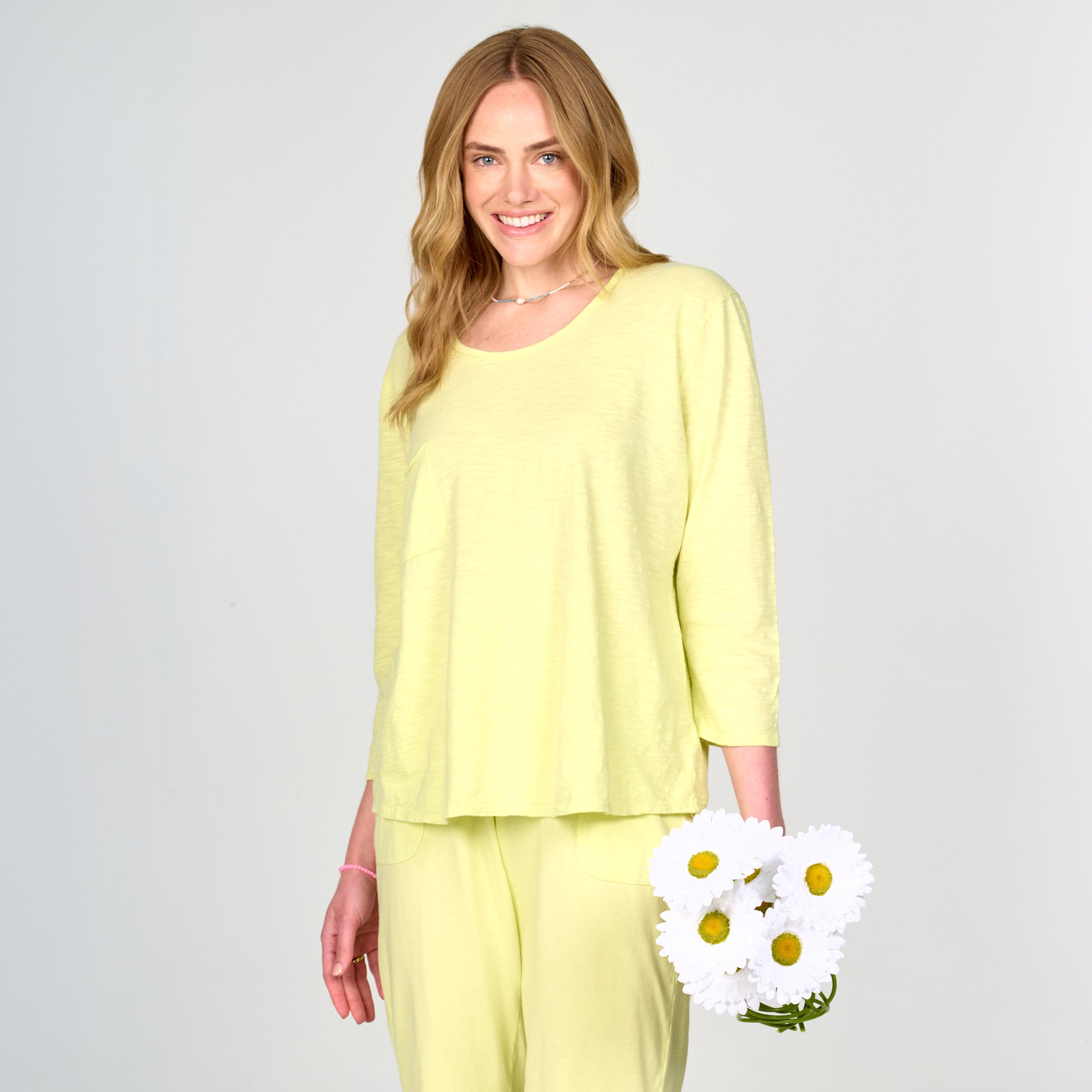 A woman with long blonde hair is smiling and holding a small bouquet of white daisies while wearing the Fresh Produce Clothes Forever Top in light yellow, pictured against a plain light background.