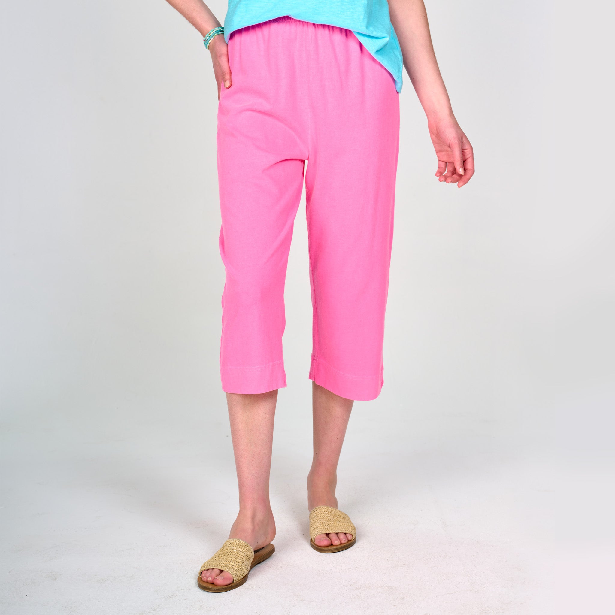 A person is wearing Fresh Produce Clothes’ bright pink Jersey Capris, featuring an elastic waistband that sits comfortably at the natural waistline. The capris are styled with a light blue top, tan woven sandals, and a bracelet. The individual is standing against a plain light background with their face not visible.