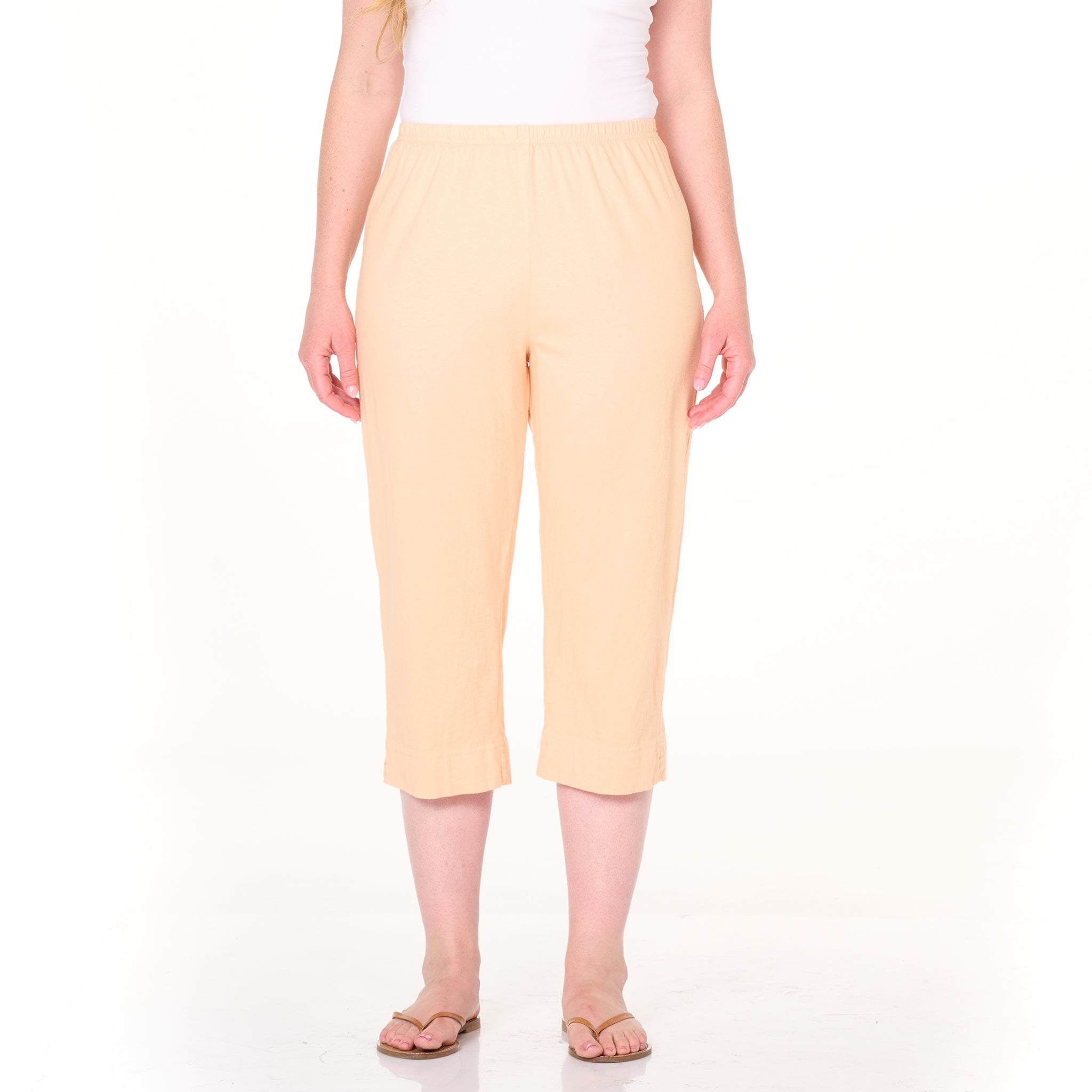 A person wearing Fresh Produce Clothes’ Jersey Capris Summer Colors in light peach, featuring an elastic waistband, paired with a white sleeveless top and beige sandals, stands on a white background. Only the lower half of the body is visible.