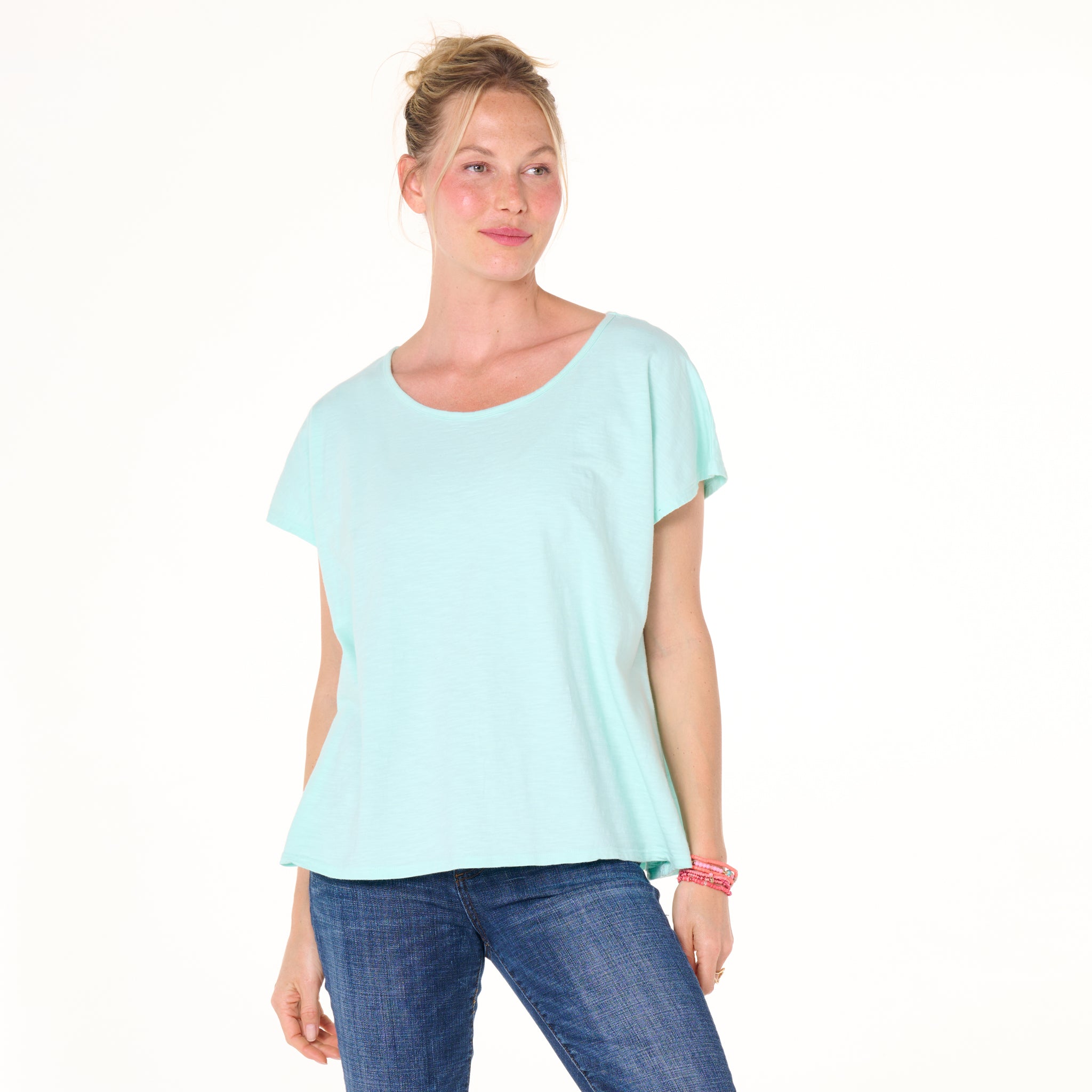 A woman with blonde hair in a loose bun wears the Fresh Produce Clothes Keepsake T- Summer Colors, a one-size, light blue t-shirt made from soft cotton slub, paired with blue jeans. She stands against a white background and smiles slightly.