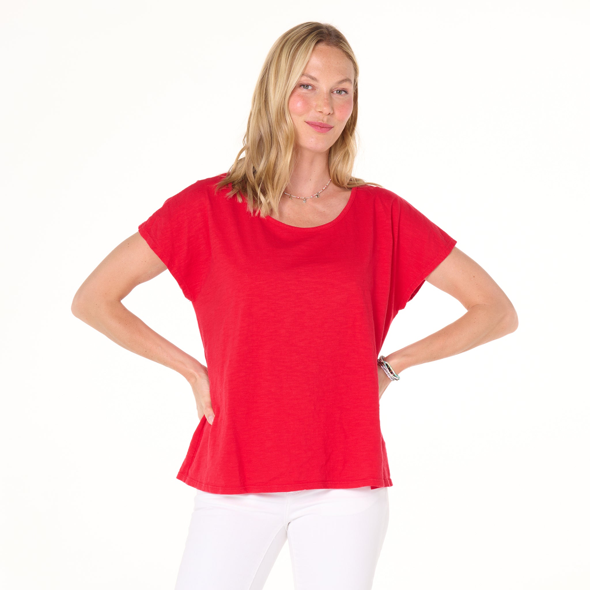 A woman with blonde hair wears the Keepsake T - 2025 Fall Colors by Fresh Produce Clothes in bright red, paired with white pants, standing with her hands on her hips and smiling against a plain white background.