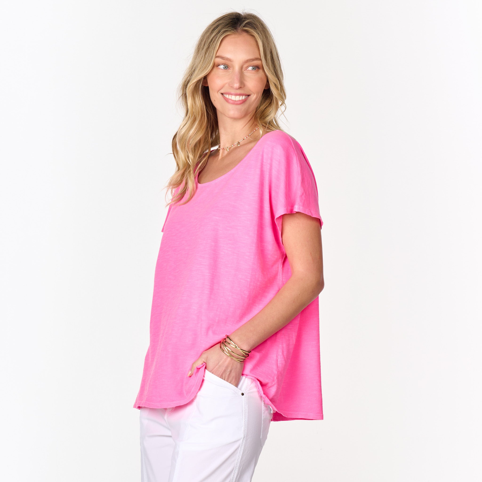 A woman with long blonde hair, dressed in the Fresh Produce Clothes Keepsake T | Clearance—a bright pink, comfortable, one-size short-sleeve tee—and white pants, stands sideways with her hand in her pocket and smiles while facing left against a white background.