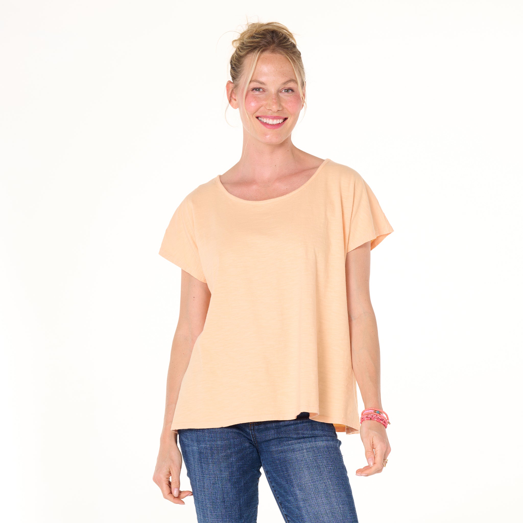 A woman with blonde hair in a bun smiles, wearing the Fresh Produce Clothes Keepsake T- Summer Colors, a peach cotton slub short-sleeve top, paired with blue jeans, standing against a plain white background.