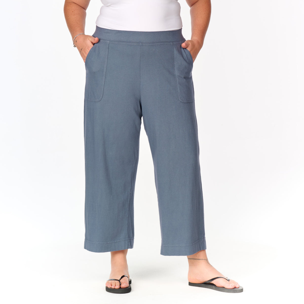 A person wearing the Fresh Produce Clothes Key Largo Capris | Plus Size—a blue, mid-rise, wide-leg cropped style—paired with a white top and black flip-flops stands with hands in pockets against a plain white background.