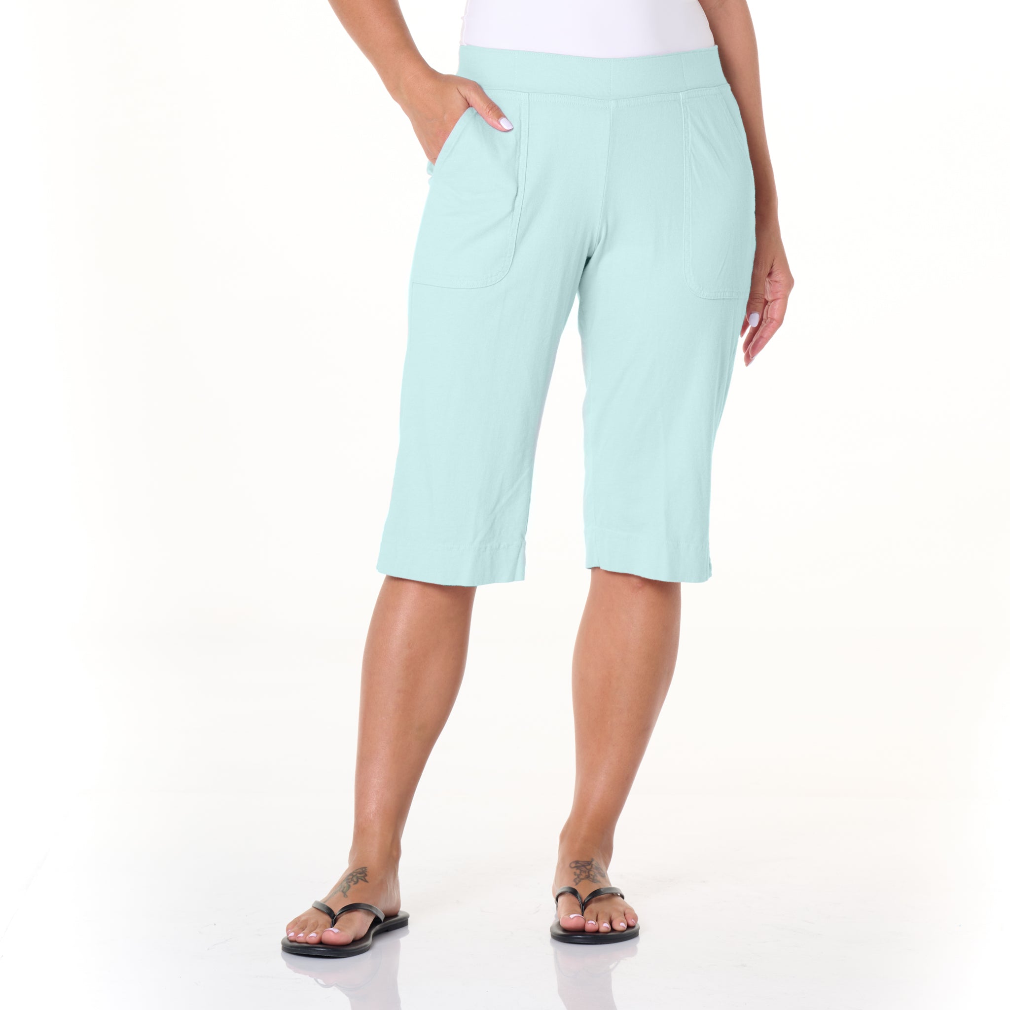 A person stands with one hand in a pocket, wearing the Fresh Produce Clothes Key Largo Pedal Pushers Summer Colors shorts in light blue, paired with a white top and black flip-flops. The background is plain white.