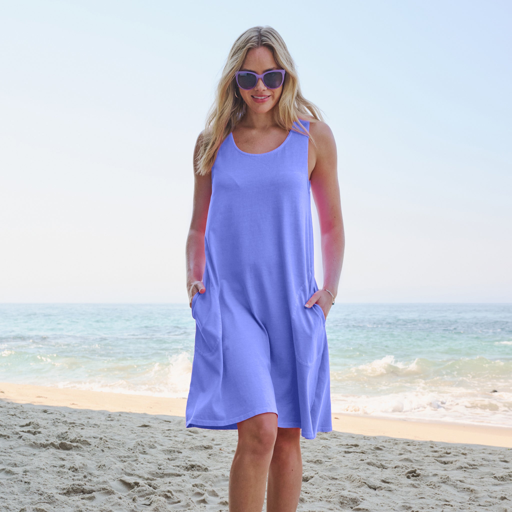Wearing the light purple La Jolla Dress by Fresh Produce Clothes and sunglasses, a woman stands on a sandy beach with gentle waves and a clear sky in the background.
