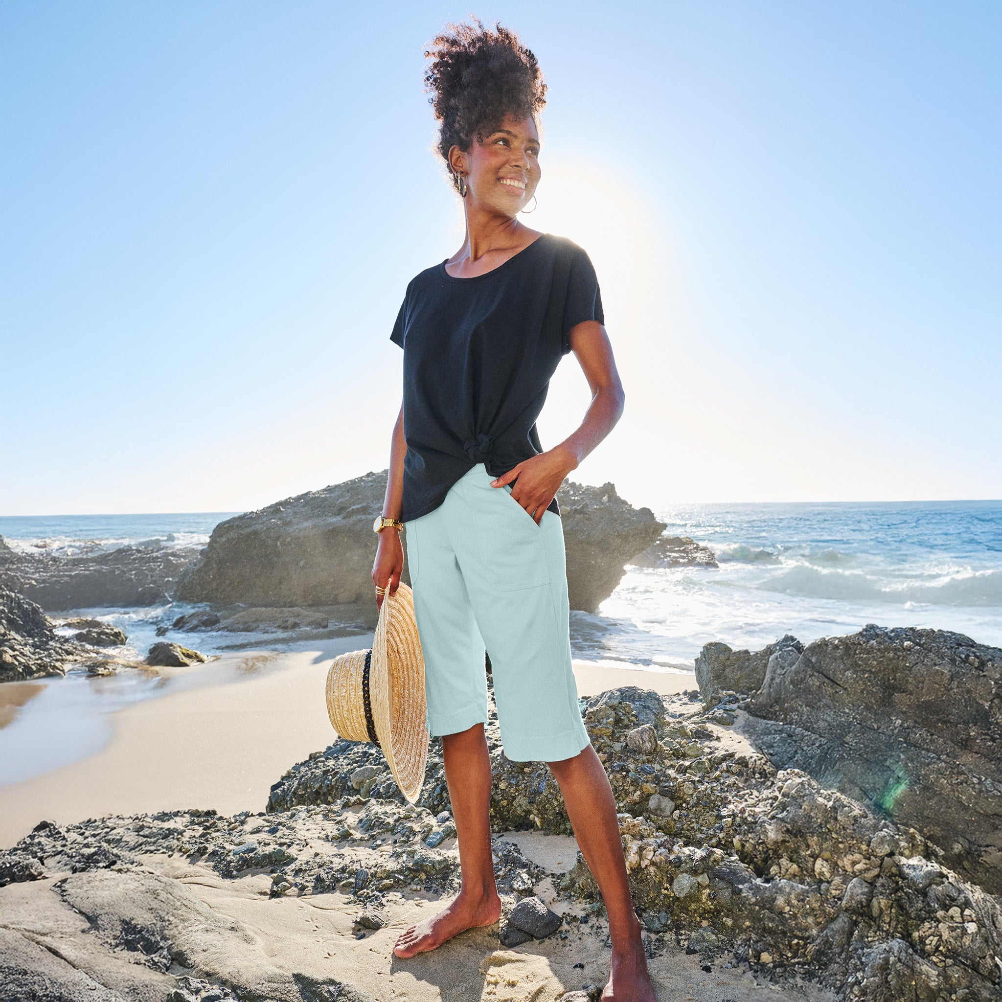 A woman stands barefoot on rocky beach sand, wearing a black cotton jersey top and Fresh Produce Clothes’ Key Largo Pedal Pushers Summer Colors, holding a straw hat, with the ocean and bright sun behind her.