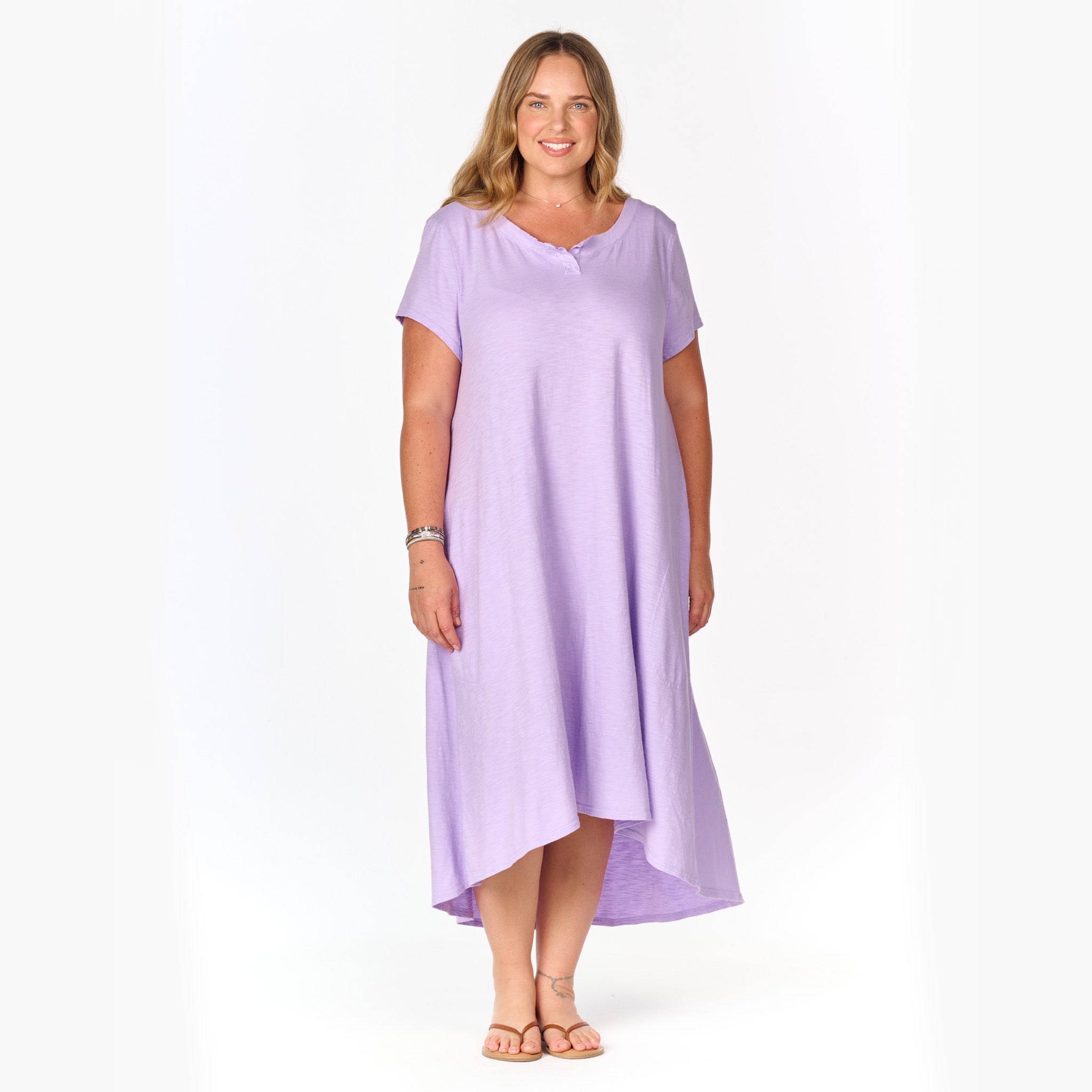 A woman stands smiling, wearing the Madeline Dress by Fresh Produce Clothes—a plus size, loose-fitting lavender midi-maxi dress with short sleeves and a high-low hemline that creates a relaxed silhouette. She pairs it with sandals and bracelets while posing against a plain white background. This style is currently on clearance.