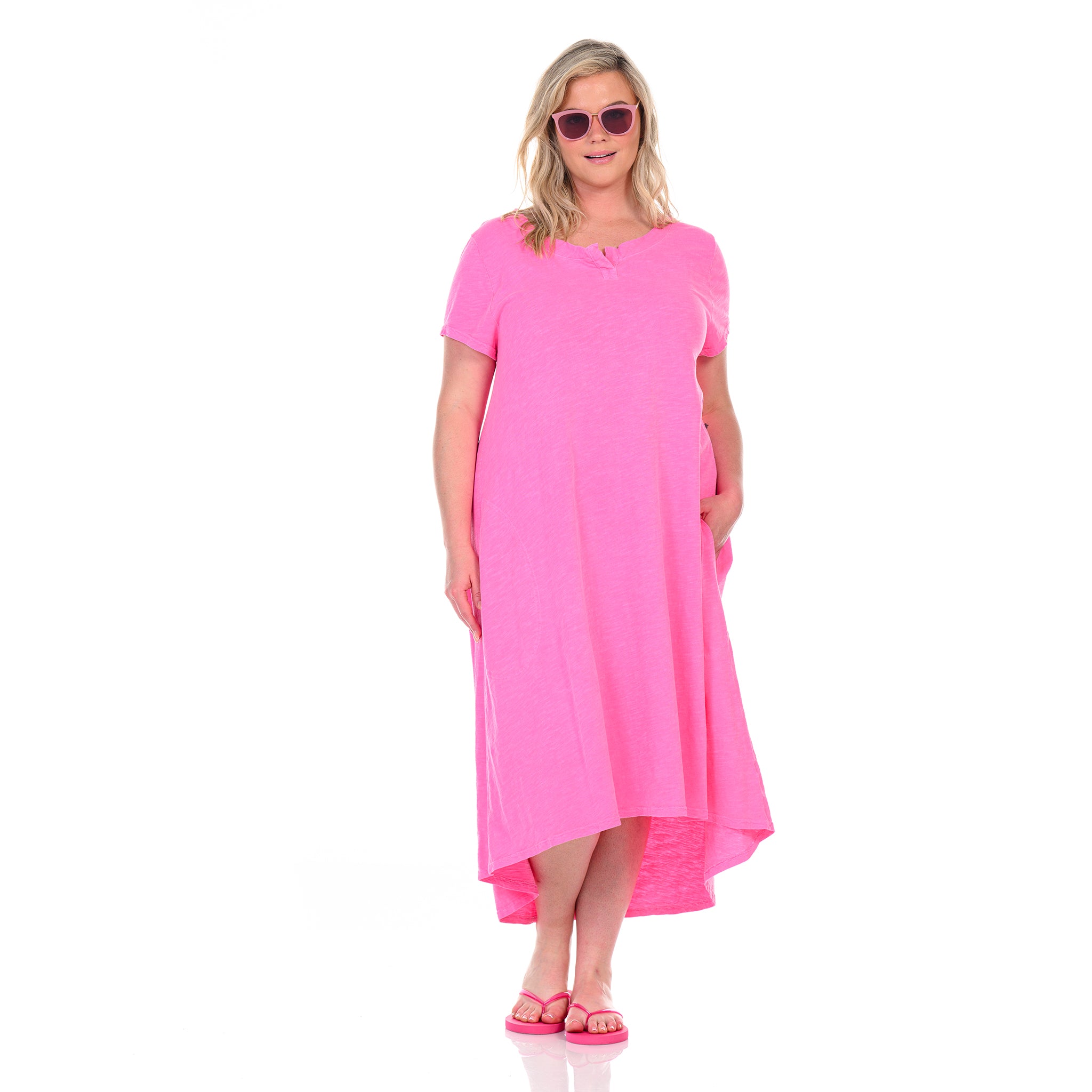 A woman wearing the Fresh Produce Clothes Madeline Dress in plus size, featuring a vibrant pink color and high-low hem with a relaxed silhouette, pairs it with pink flip-flops and matching pink sunglasses as she poses against a white background, smiling with one hand in her pocket.