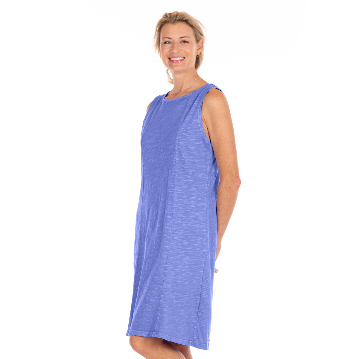 A woman stands smiling against a plain white background, wearing the Marissa Dress by Fresh Produce Clothes—a sleeveless, knee-length blue A-line dress.