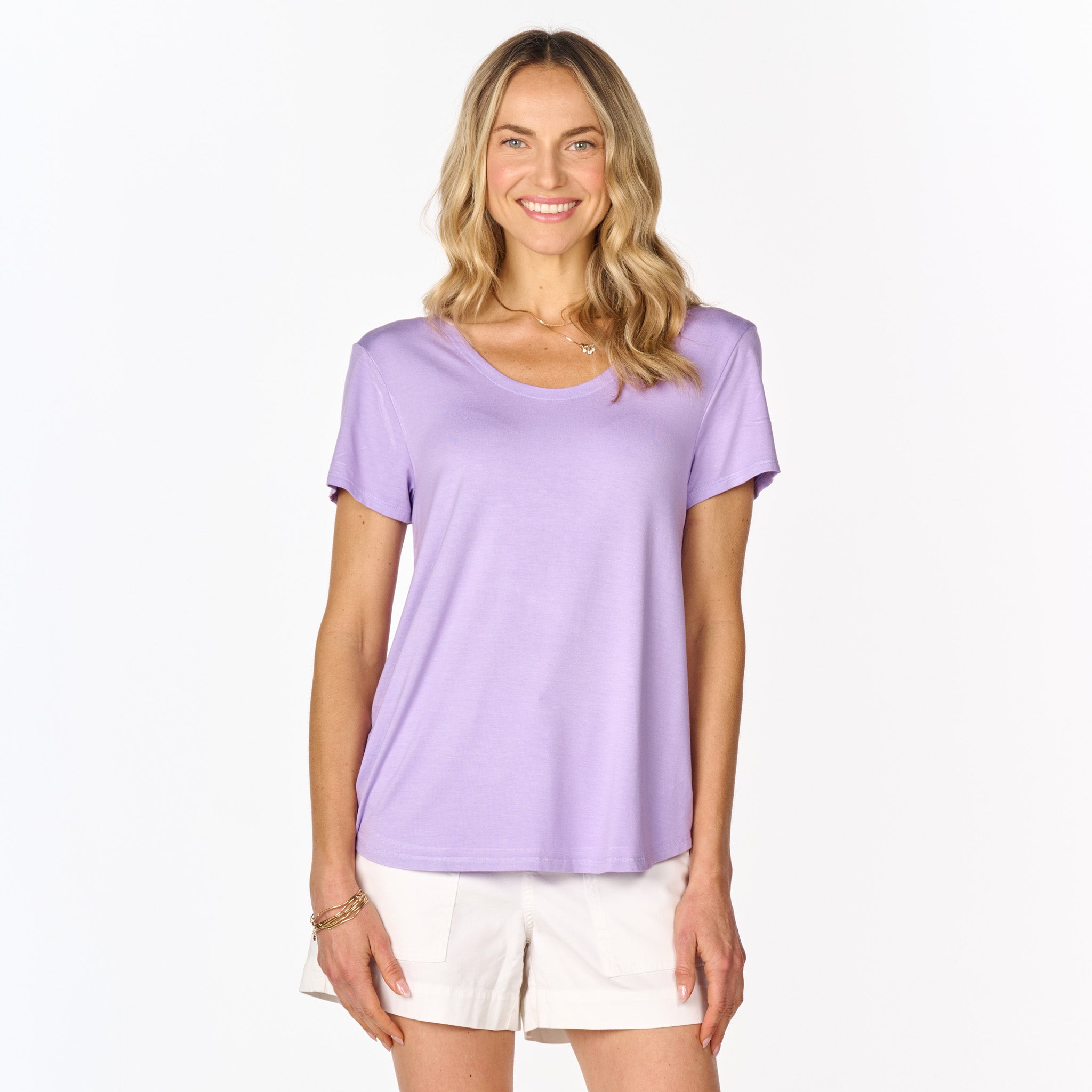 A smiling woman with long blonde hair wears the Mika T from Fresh Produce Clothes, a lavender rayon lycra top with a twist back detail, paired with white shorts and standing against a plain white background.
