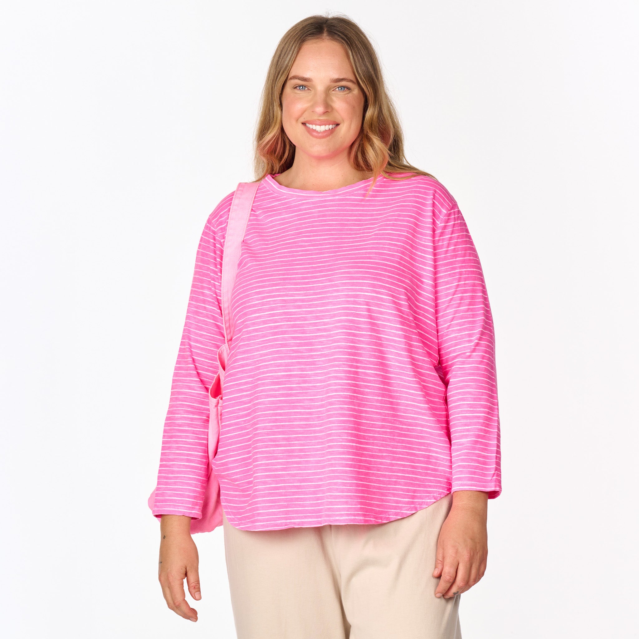 A smiling person with long hair is wearing the Fresh Produce Clothes Ocean Stripe Catalina Top (Plus Size, Clearance), a loose-fitting cotton 3/4 sleeve top in the Ocean Stripe print, paired with beige pants and carrying a pink bag over one shoulder while standing against a white background.