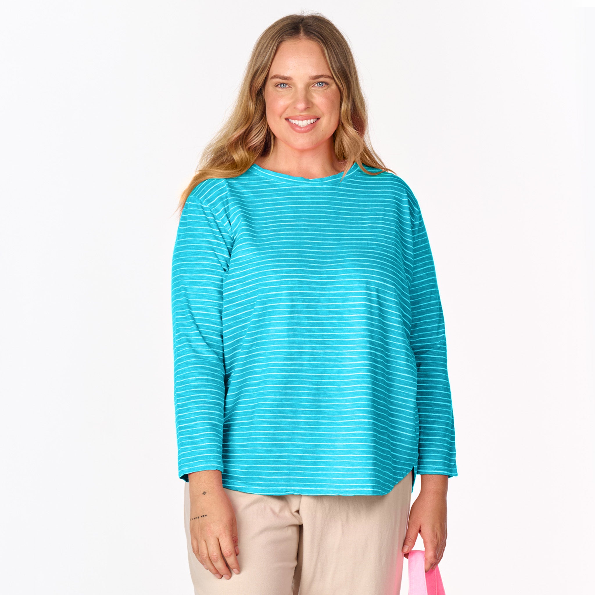 A woman with long, wavy hair wearing the Fresh Produce Clothes Ocean Stripe Catalina Top (Plus Size, Clearance) and beige pants smiles while holding a pink bag.