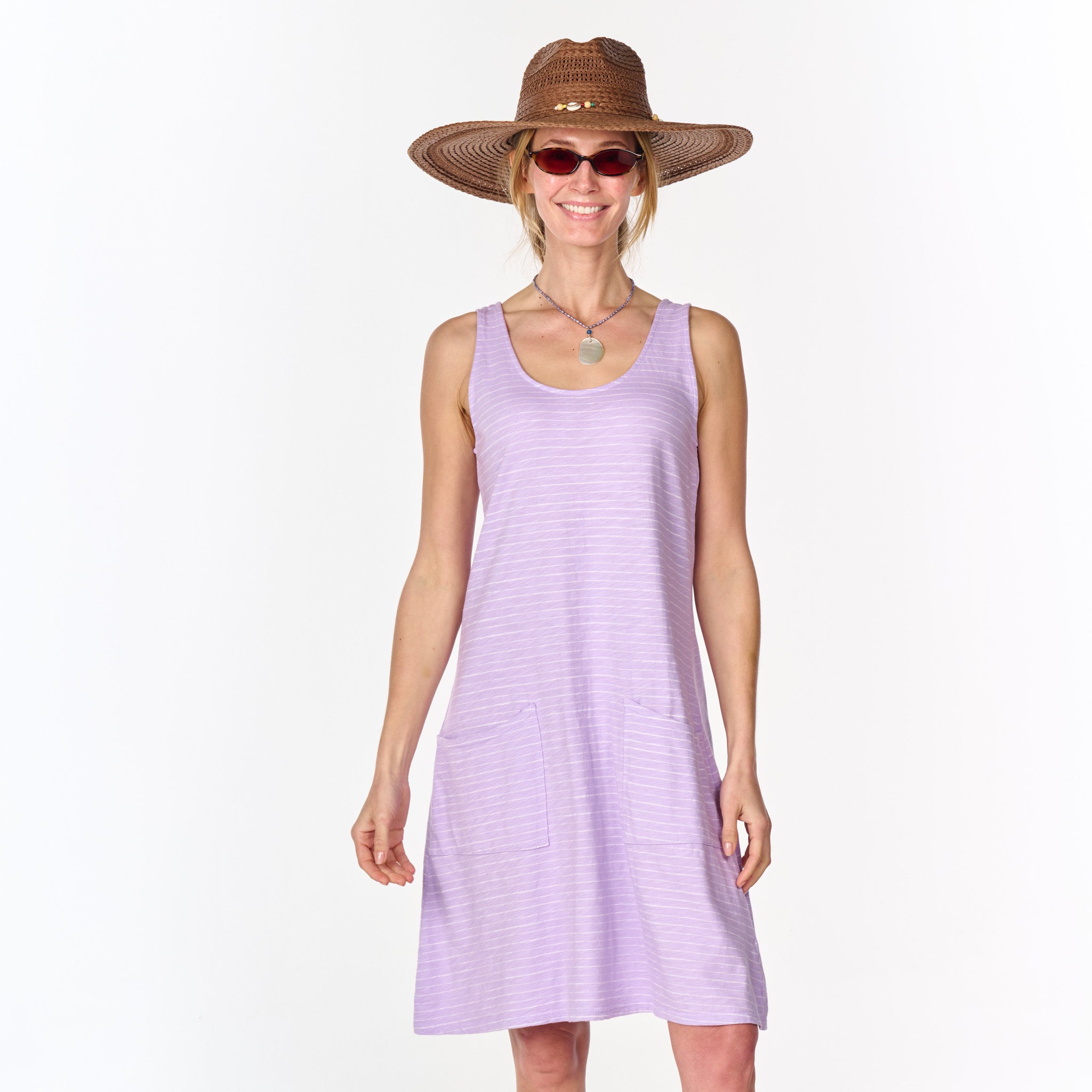 A woman wearing a wide-brimmed straw hat, sunglasses, a necklace, and the Fresh Produce Clothes Ocean Stripe Drape Dress in light purple cotton slub with pockets stands smiling against a plain white background.