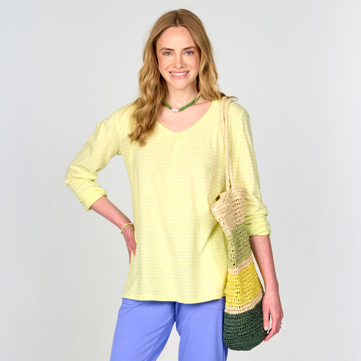 A woman with long blonde hair smiles, dressed in a light yellow long-sleeve top, blue pants, and a green-yellow crochet shoulder bag. Against a simple pale background, her easygoing style brings to mind the Ocean Stripe Hoodie by Fresh Produce Clothes—a lightweight cotton hoodie made in the USA, now available on clearance.