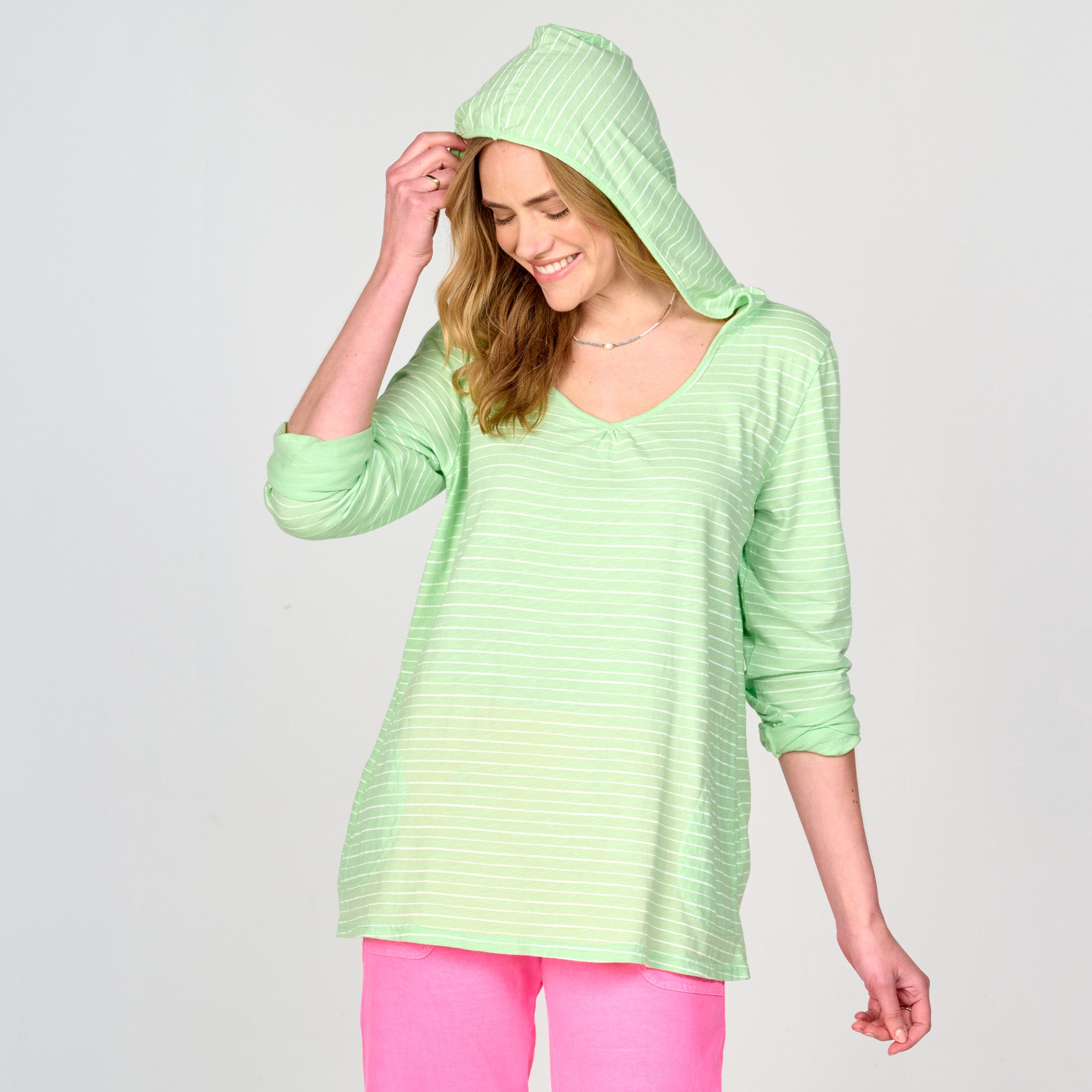 A woman smiles while wearing the Fresh Produce Clothes Ocean Stripe Hoodie in a light green novelty stripe print, paired with bright pink pants and the hood up, standing against a plain light gray background. This cozy hoodie also features hidden side pockets for added convenience.
