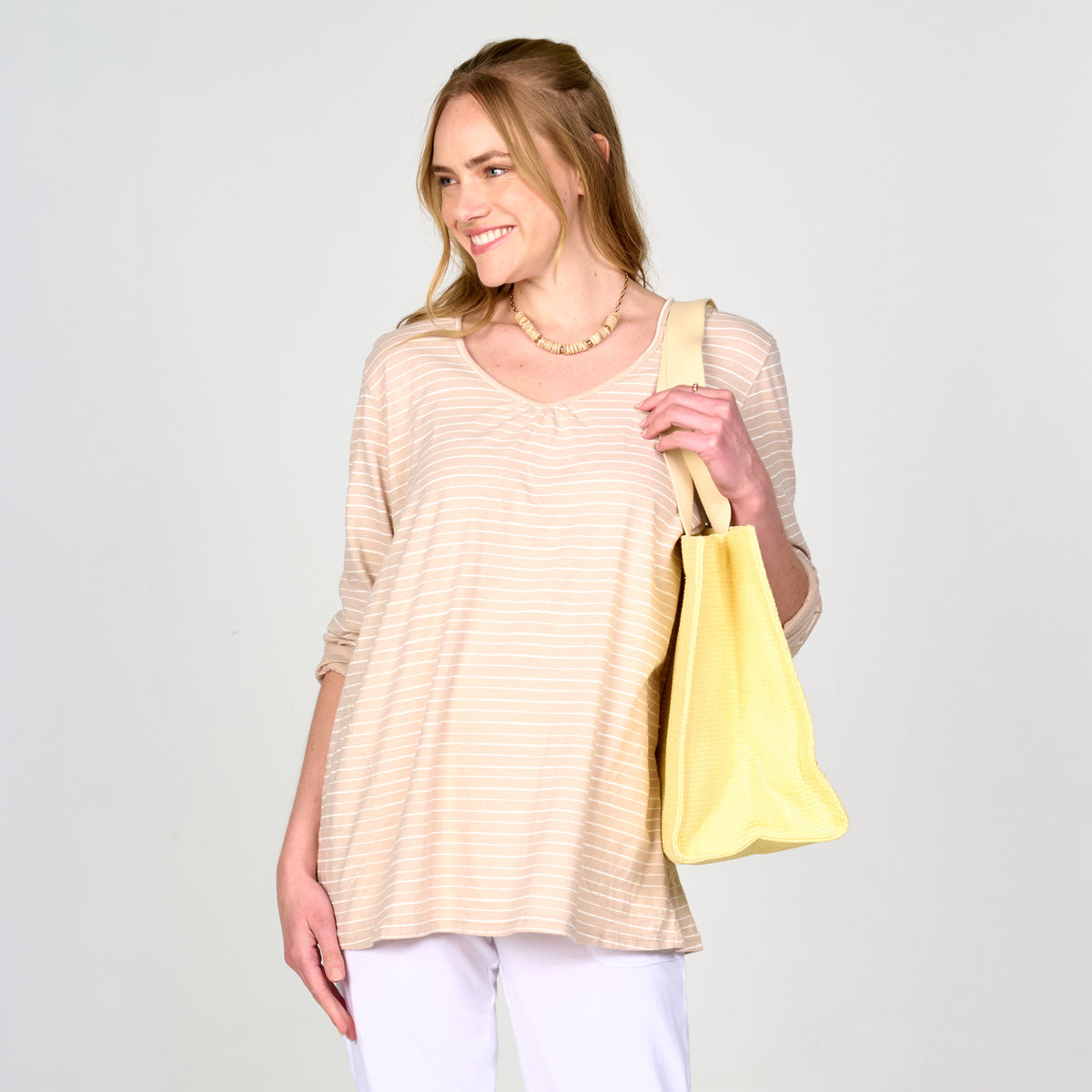 A woman with long blonde hair smiles as she poses against a simple light background, wearing the Ocean Stripe Hoodie by Fresh Produce Clothes in beige stripes, layered over a top and paired with white pants. She accessorizes with a beaded necklace and carries a large yellow tote bag over her shoulder.