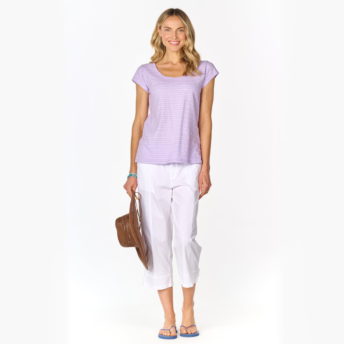 Smiling woman wearing a light purple Ocean Stripe Vintage Scoop Neck T by Fresh Produce Clothes, which features a slimming shape, paired with white cropped pants and sandals, holding a brown woven handbag, and standing against a white background.