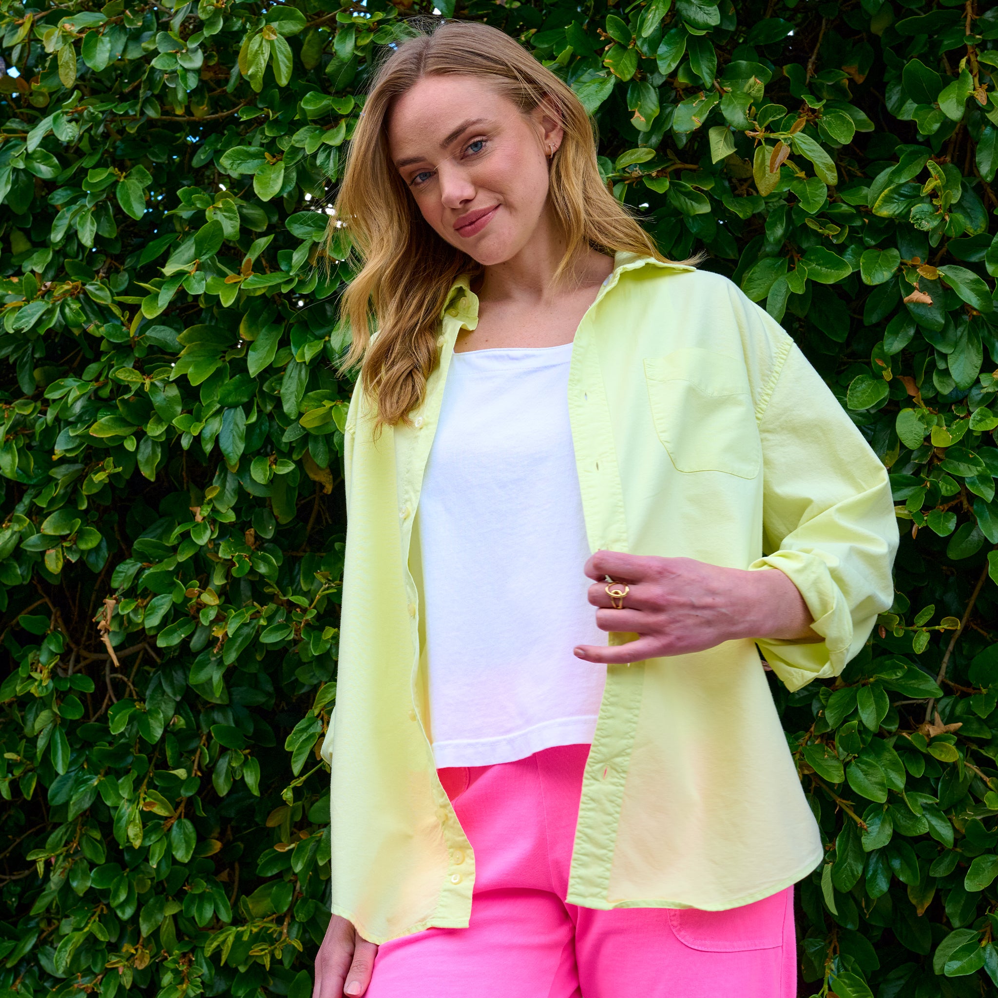 A woman stands in front of green leaves, smiling softly at the camera, wearing a relaxed fit light yellow Classic Oxford Shirt from Fresh Produce Clothes over a white top and pink pants.
