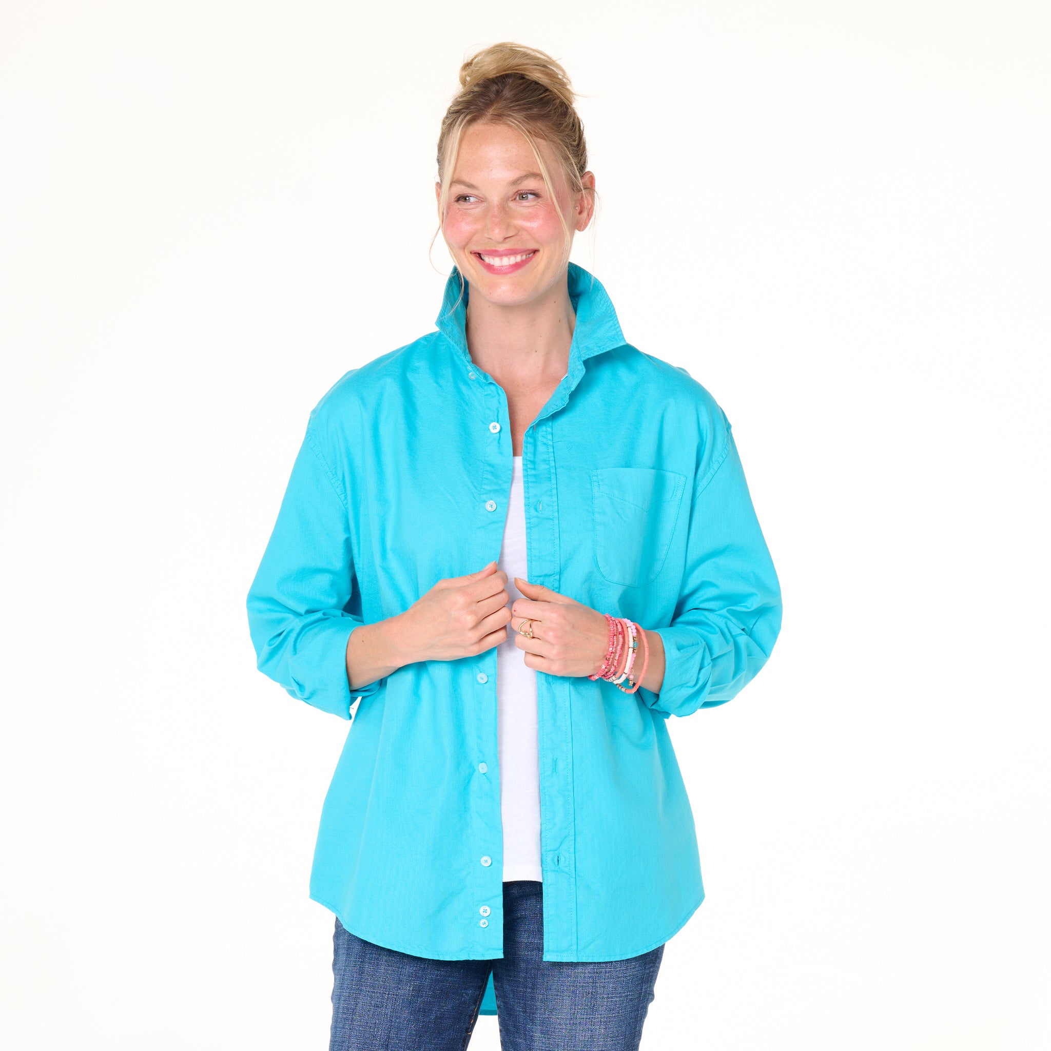 A smiling woman with blonde hair in a bun wears the Fresh Produce Clothes Classic Oxford Shirt in bright turquoise—styled relaxed over a white top and jeans—standing against a plain white background.