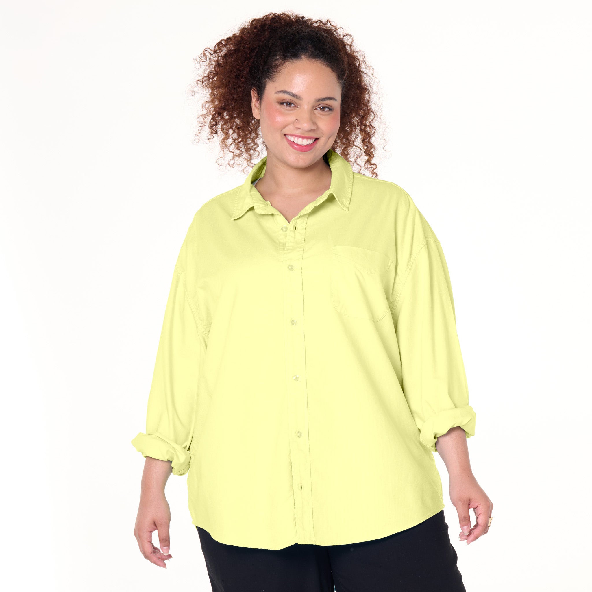 A smiling woman with curly hair wears the Classic Oxford Shirt in light yellow from Fresh Produce Clothes, styled with rolled sleeves and paired with black pants, standing against a plain white background. This plus size shirt is currently on clearance.