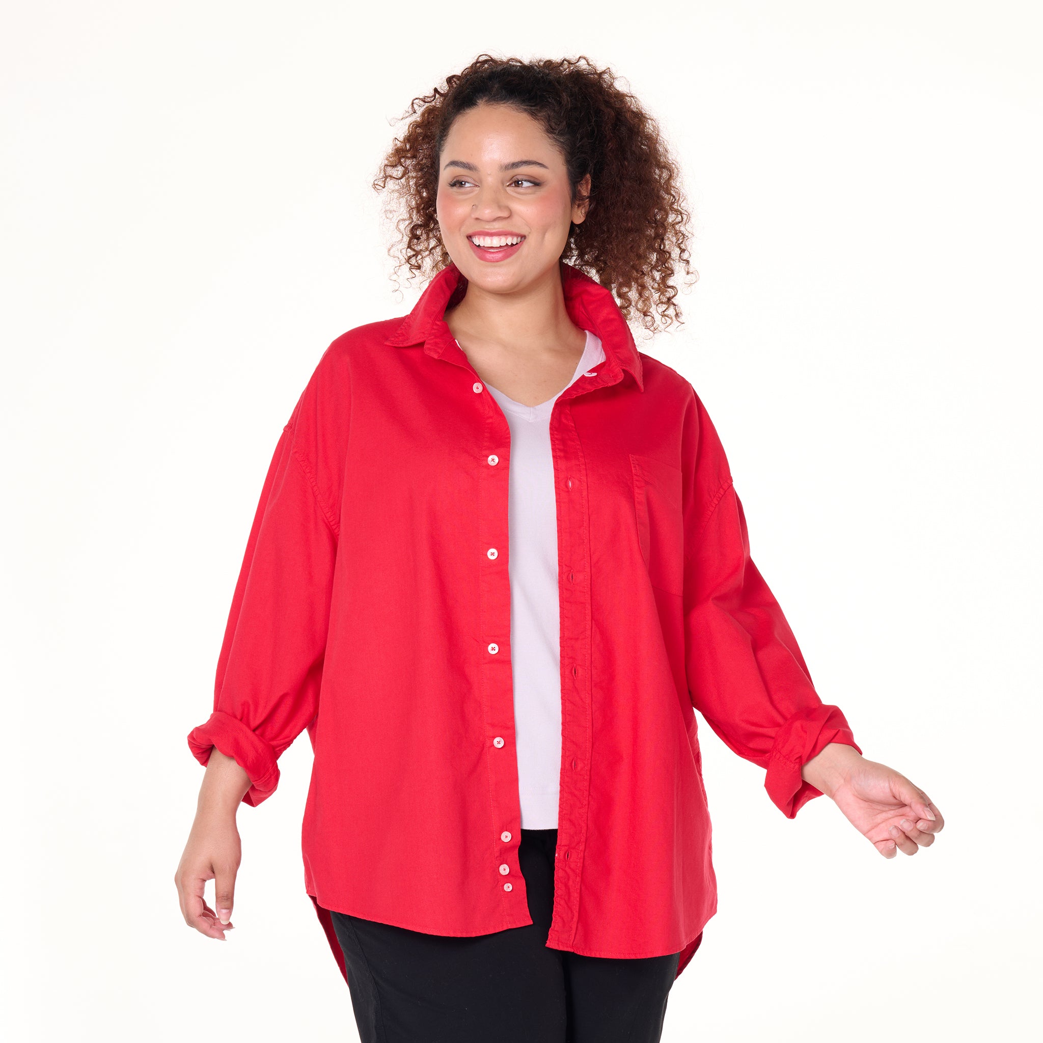 A smiling woman with curly hair models the Fresh Produce Clothes Classic Oxford Shirt in bright red—part of the Plus Size Clearance collection—styled over a white top and black pants, standing against a plain white background.