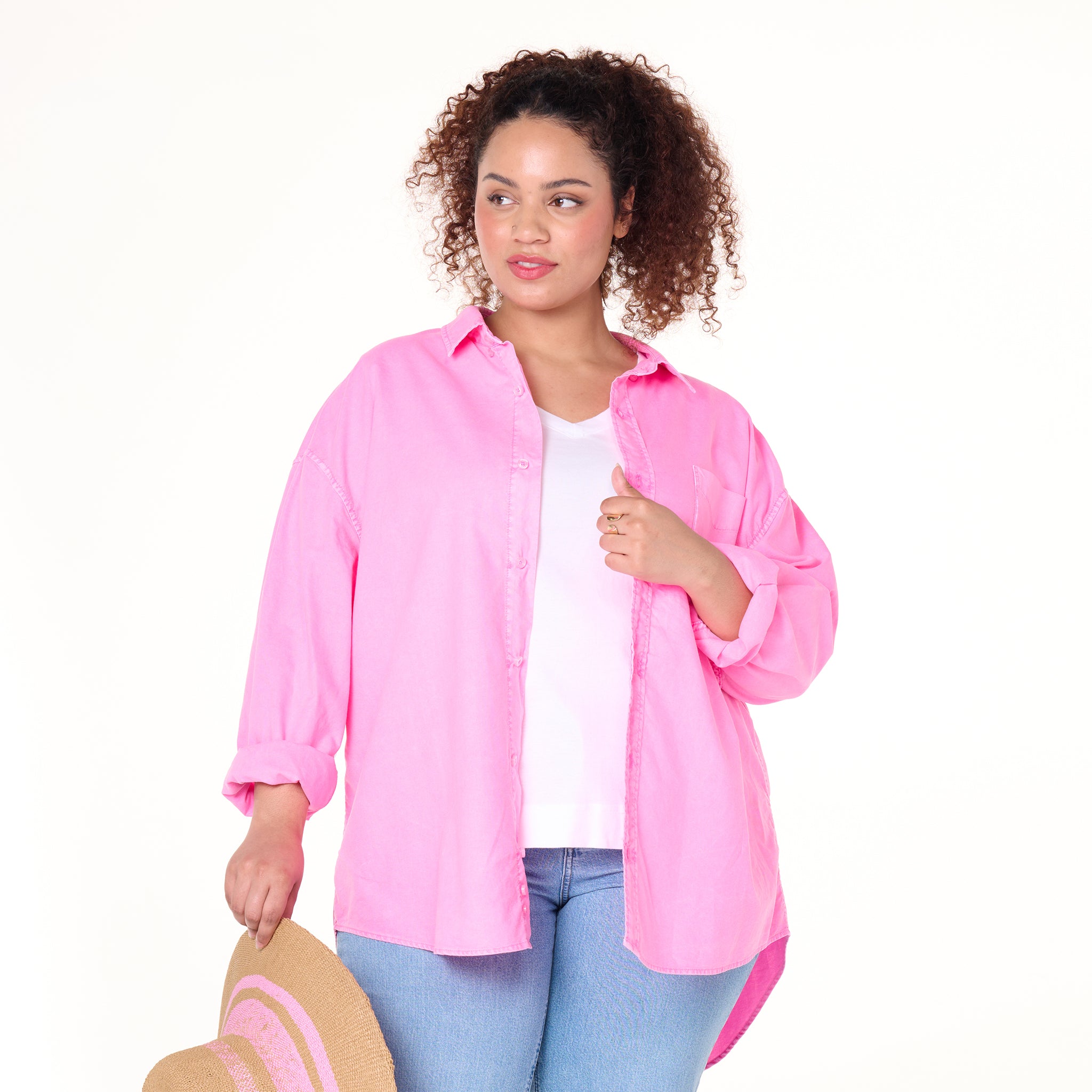 A woman with curly hair models the Classic Oxford Shirt in bright pink from Fresh Produce Clothes, styled oversized over a white top and blue jeans. She holds a straw hat with pink accents, gazing to the side against a plain white background. The shirt is available in plus size and currently on clearance.