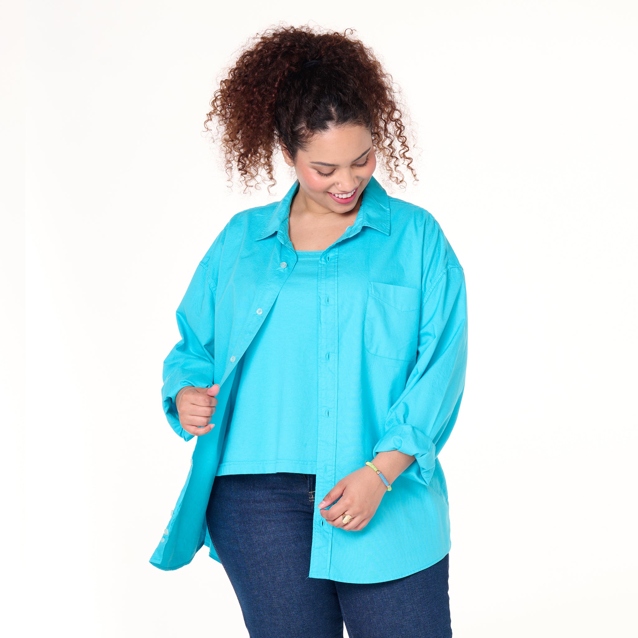 A woman with curly hair, smiling and looking down, wears the Fresh Produce Clothes Classic Oxford Shirt | Plus Size | Clearance in bright turquoise over a matching top and dark jeans, holding the shirt open with one hand against a plain white background.