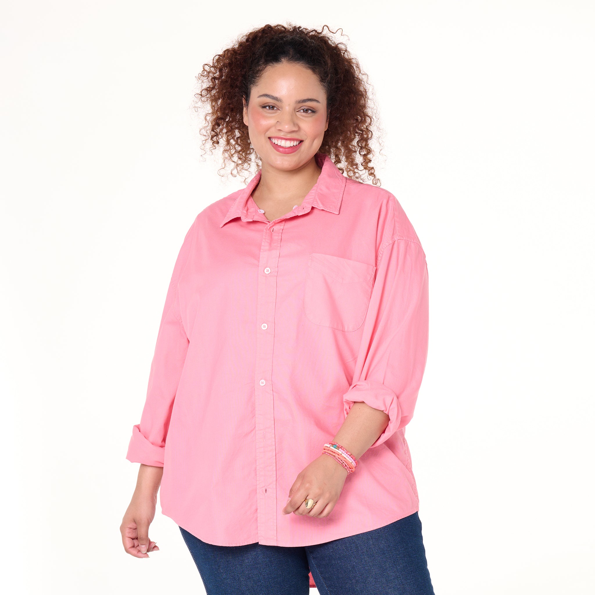 A woman with curly hair smiles while wearing the Fresh Produce Clothes Classic Oxford Shirt in pink, available in plus sizes and on clearance, with rolled sleeves and blue jeans, standing against a white background.