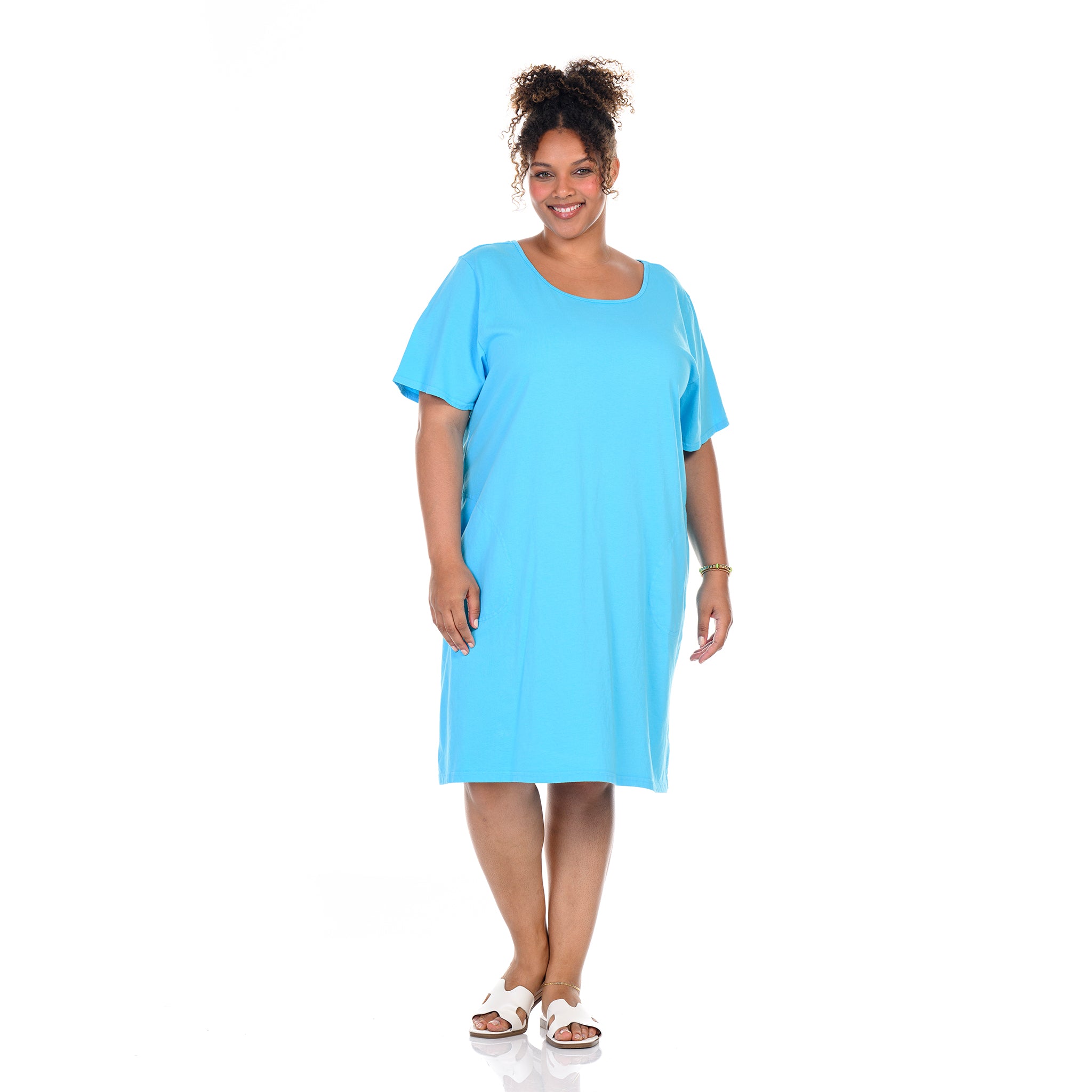 A woman smiles while standing on a white background, wearing the Fresh Produce Clothes Sanibel Dress | Plus Size Blue Fin—a loose-fitting, short-sleeve, knee-length bright blue cotton jersey t-shirt dress—paired with white sandals.