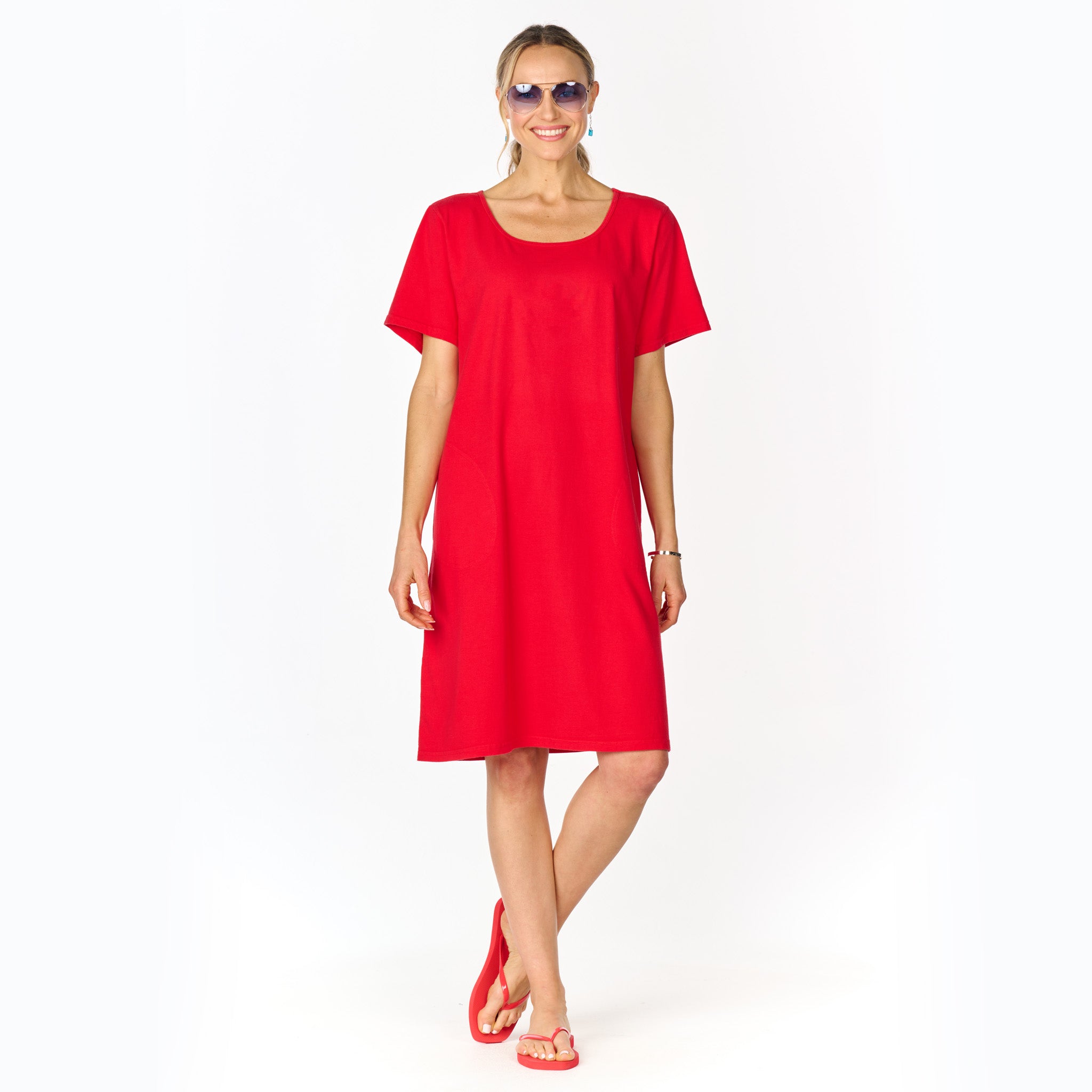 A smiling woman wearing sunglasses, the Sanibel Dress in plain red cotton jersey with short sleeves by Fresh Produce Clothes, and red sandals stands against a white background.