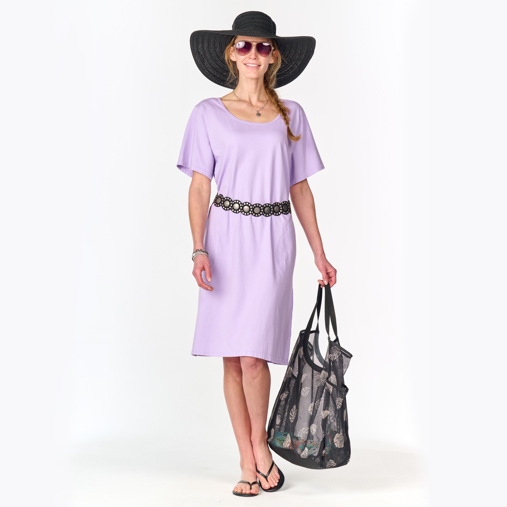 A woman wearing the Sanibel Dress by Fresh Produce Clothes, styled as an oversized lavender cotton jersey t-shirt dress, paired with a black sunhat, sunglasses, and sandals, carrying a large black bag and smiling while standing against a white background.