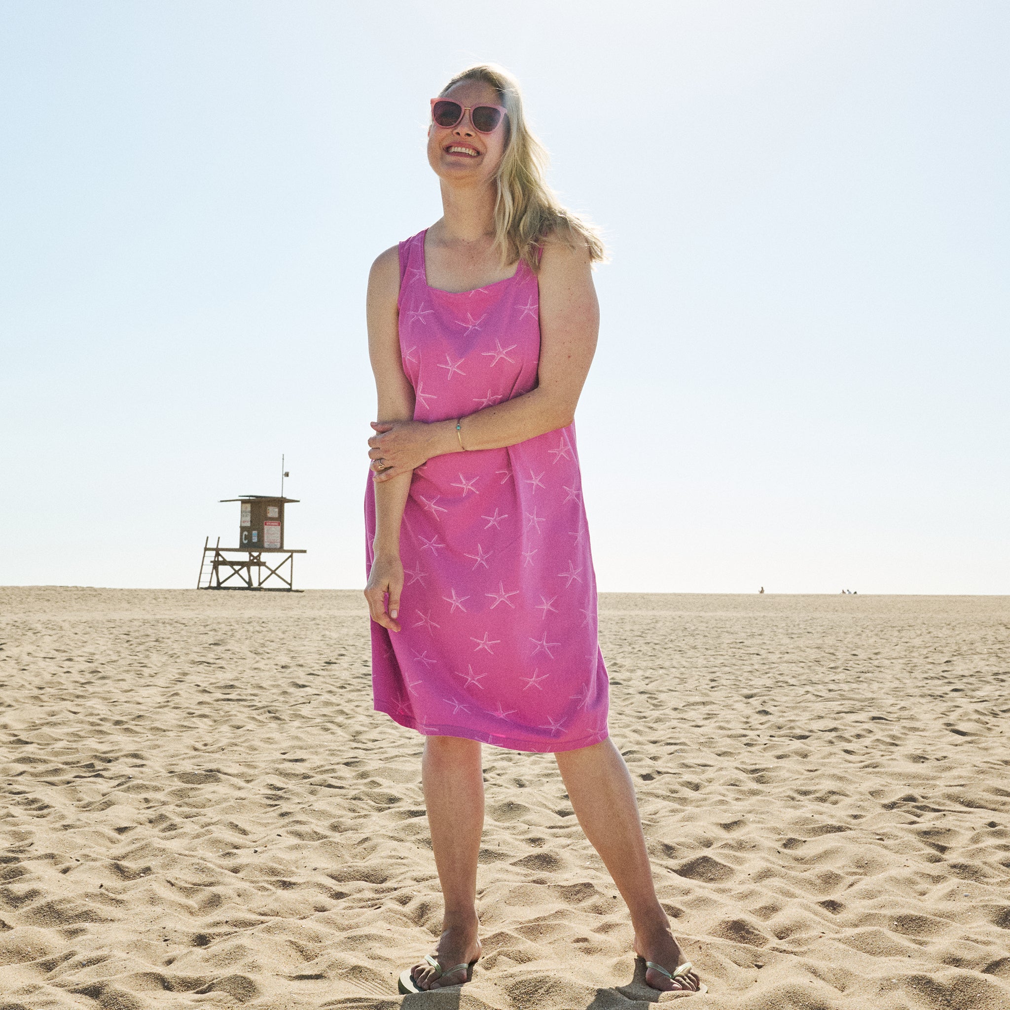 A woman in the Fresh Produce Clothes Sea Star Square Neck Dress | Plus Size CHB SUF and sunglasses stands smiling on a sandy beach, with a lifeguard tower in the background under a clear sky.