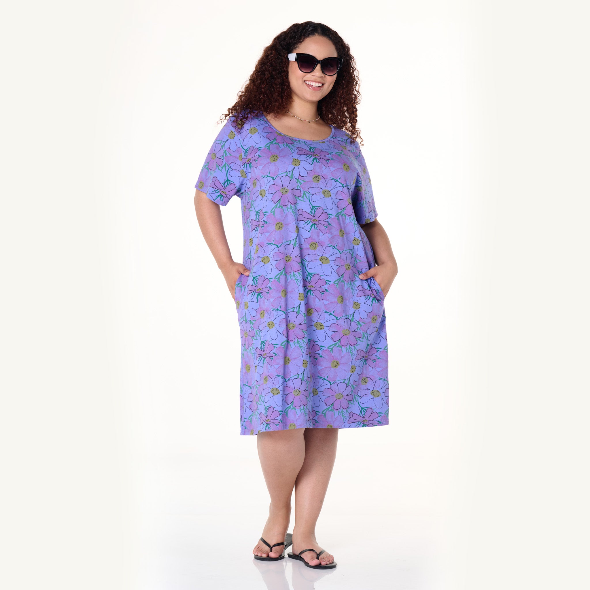 A woman wearing sunglasses and the Fresh Produce Clothes Spring Day Sanibel Dress in a comfortable, knee-length purple floral print style stands smiling with hands in pockets against a plain white background, paired with flip-flops.