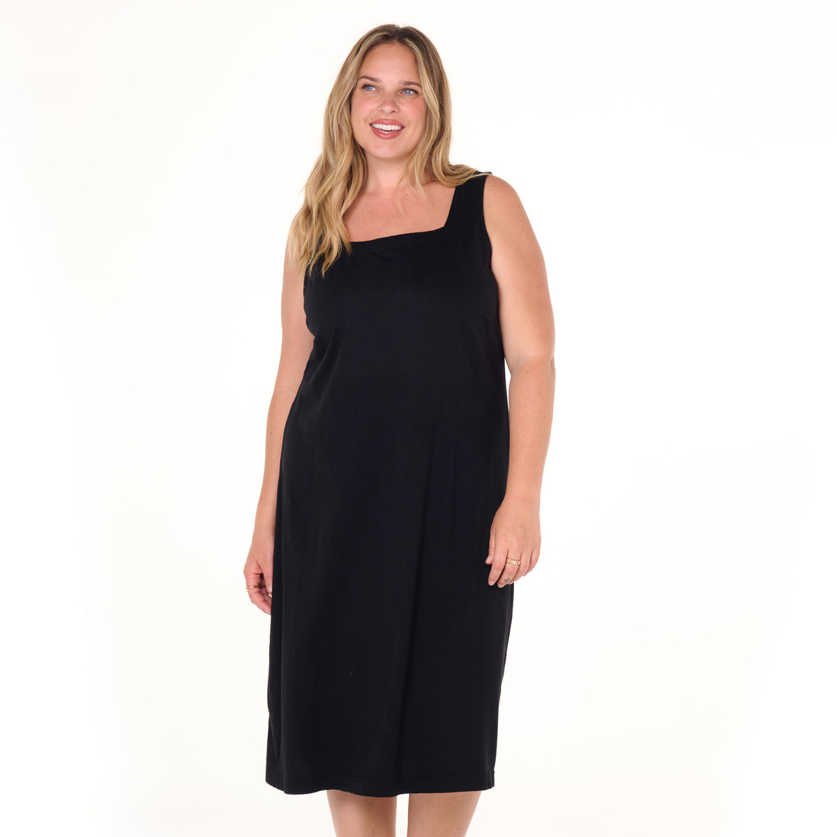 A smiling woman with long blonde hair wears the Fresh Produce Clothes Square Neck Dress | Plus Size, a sleeveless, knee-length black shift dress, standing against a plain white background.