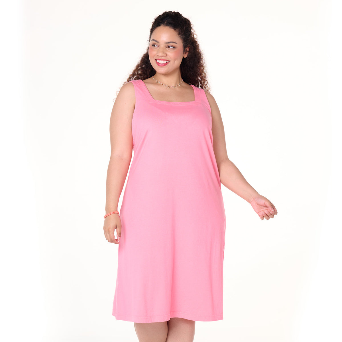 A woman with curly hair is smiling against a plain white background, wearing the Fresh Produce Clothes Square Neck Dress | Plus Size—a sleeveless, knee-length pink dress.