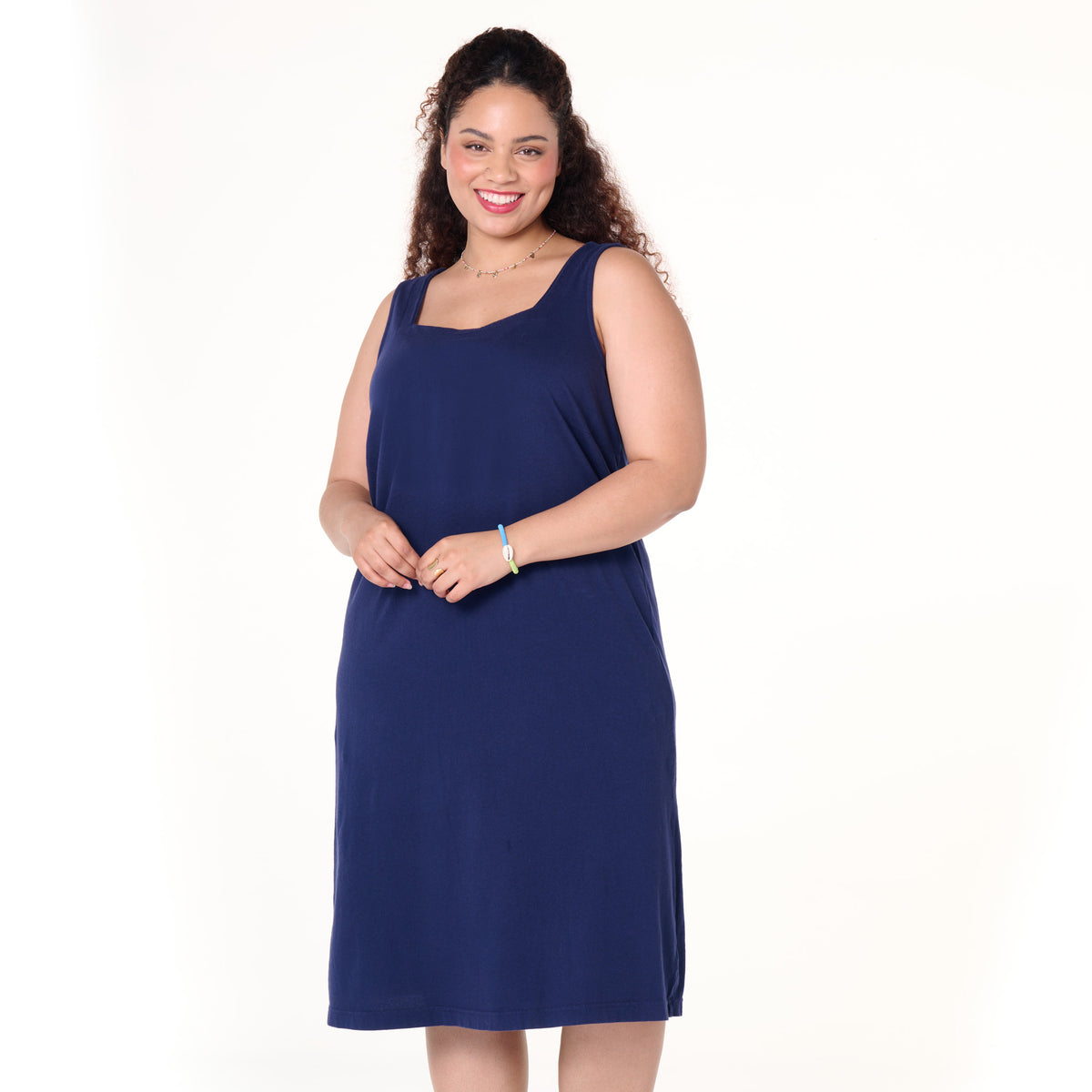 A woman with curly hair smiles, wearing the Fresh Produce Clothes Square Neck Dress | Plus Size—a sleeveless, knee-length navy blue shift dress. She stands against a plain white background, holding her hands together in front of her.