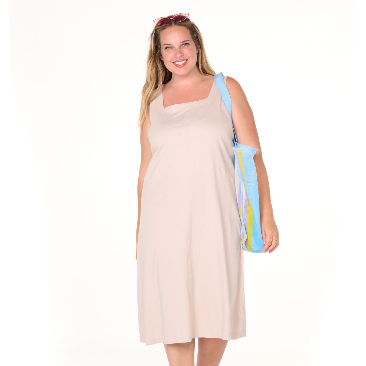 A smiling woman stands against a white background wearing the Fresh Produce Clothes Square Neck Dress | Plus Size—a sleeveless beige cotton jersey shift dress—with sunglasses on her head and carrying a blue and yellow tote bag.